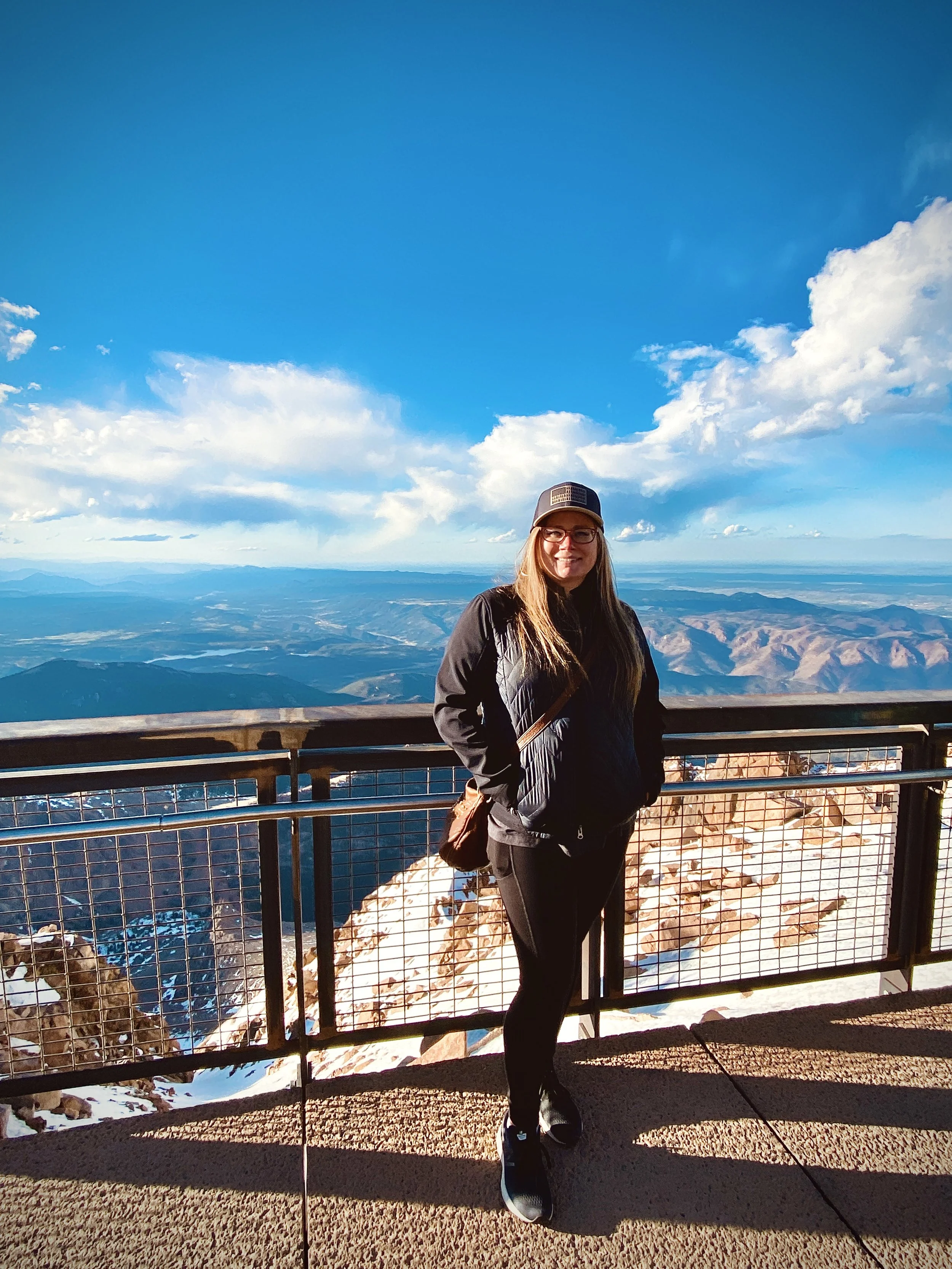 woman with long blonde hair and a hat standing in front of a railing before a wide, mountain landscape