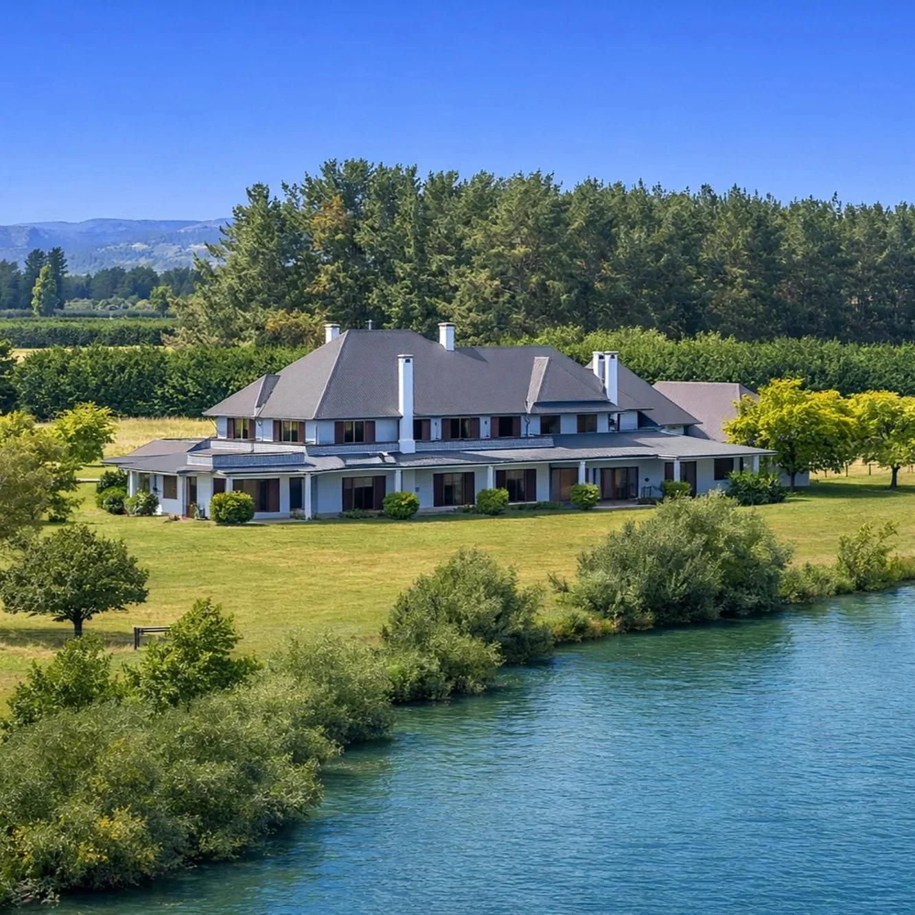 A large white house with multiple chimneys and a grey roof sits on a green lawn beside a river, surrounded by trees and distant hills under a blue sky.
