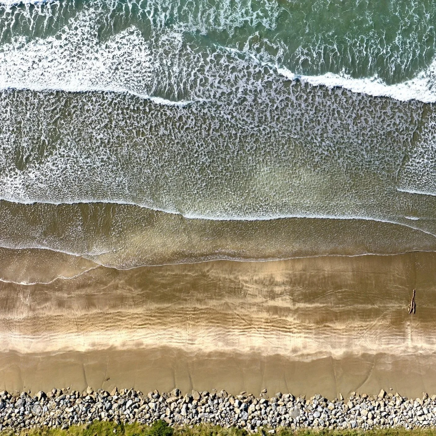 An aerial view of a beach with green ocean waves crashing onto the sandy shore, with rocks at the bottom edge of the image.