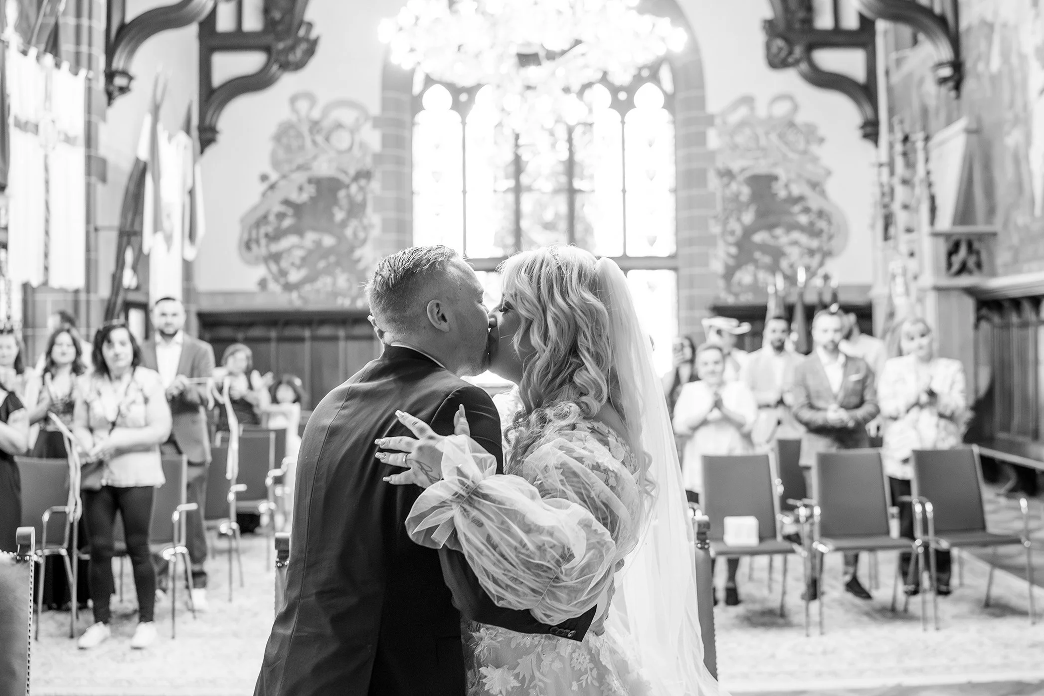 Braut und Bräutigam küssen sich während ihrer Hochzeitszeremonie in einer Schlosskapelle, mit Gästen im Hintergrund.