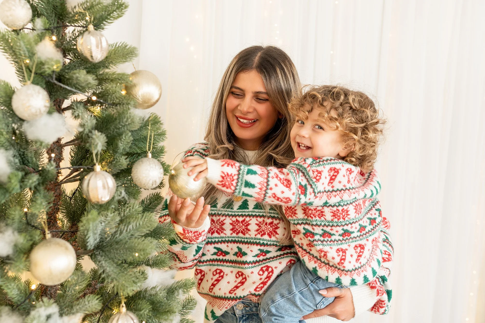 Eine Frau und ein Kind schmücken einen Weihnachtsbaum mit goldenen Kugeln, beide tragen weihnachtliche Pullover mit roten, grünen und weißen Mustern, die Frau lächelt, das Kind freut sich beim Aufhängen einer Kugel.