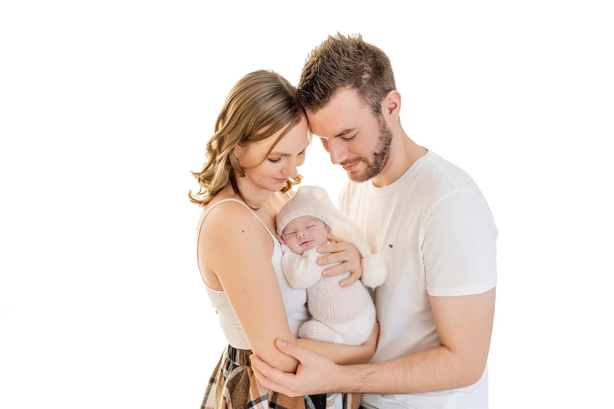 Ein Paar hält sein neugeborenes Baby, das einen Strickmütze trägt, liebevoll im Arm, beide schauen auf das Baby.