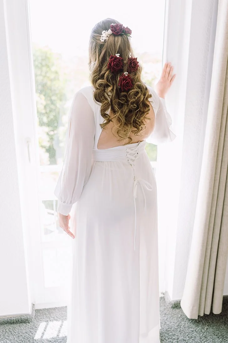 Frau in einem weißen Hochzeitskleid mit offenen Rücken, Haarkranz aus roten Rosen und kleinen weißen Blumen, schaut aus einem Fenster.