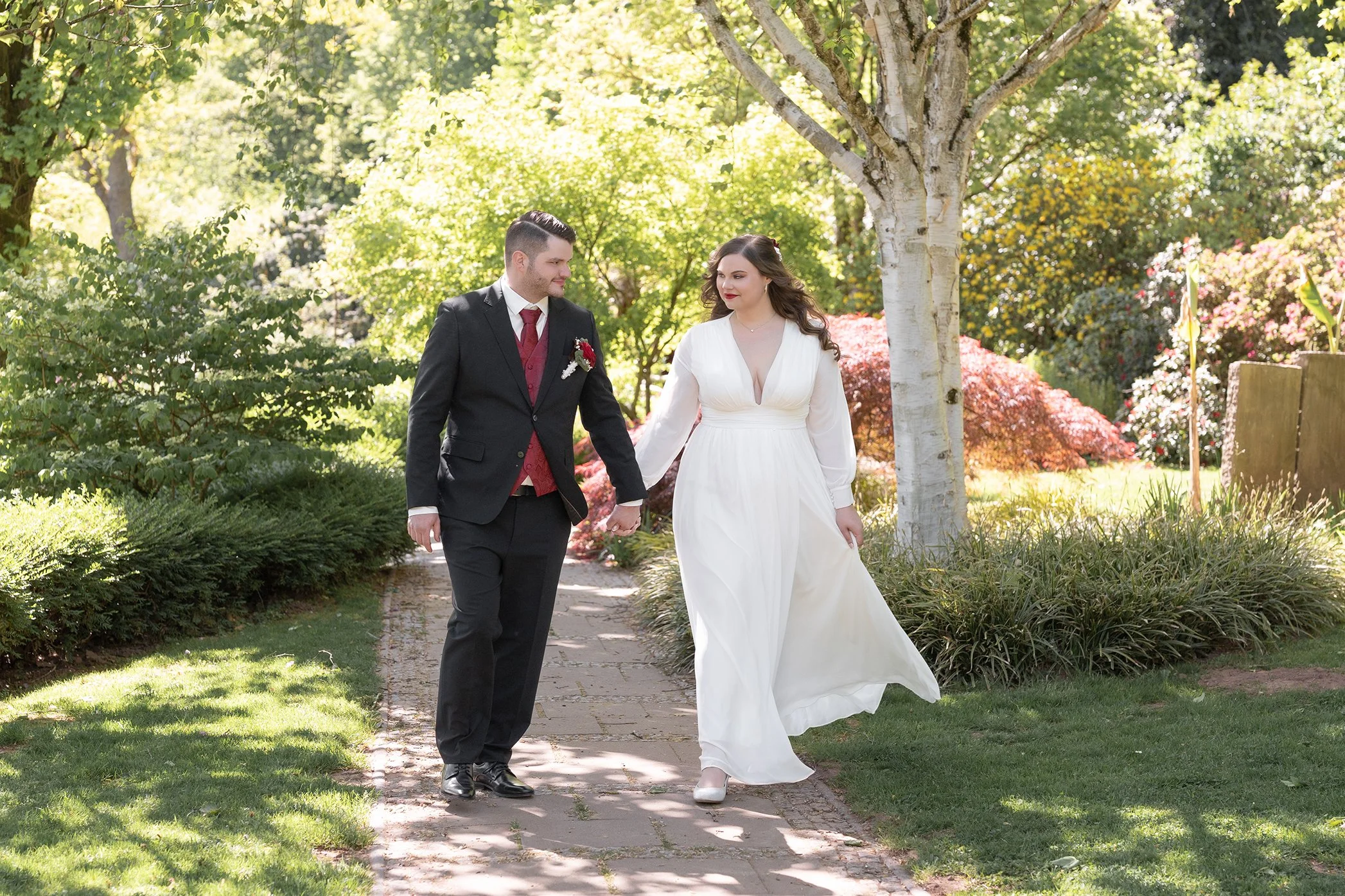 Ein Paar bei ihrer Hochzeit, spazierend durch einen Garten mit grünen Bäumen und Sträuchern, er trägt einen schwarzen Anzug mit roter Krawatte, sie trägt ein weißes Brautkleid, beide lächeln.