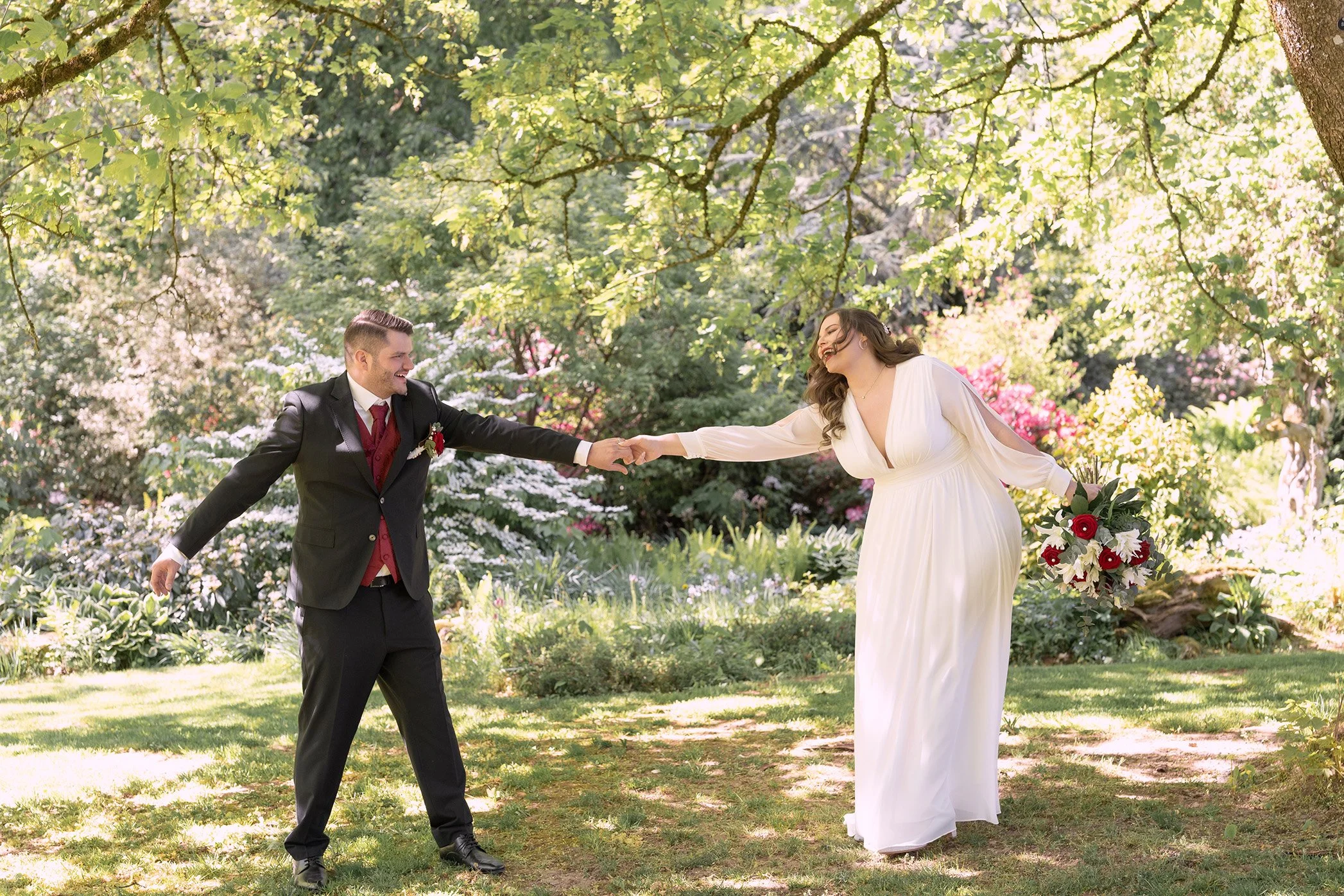 Ein Brautpaar tanzt im Garten unter einem Baum, die Frau trägt ein weißes Brautkleid und hält einen Bouquet, der Mann trägt einen schwarzen Anzug mit roter Krawatte.