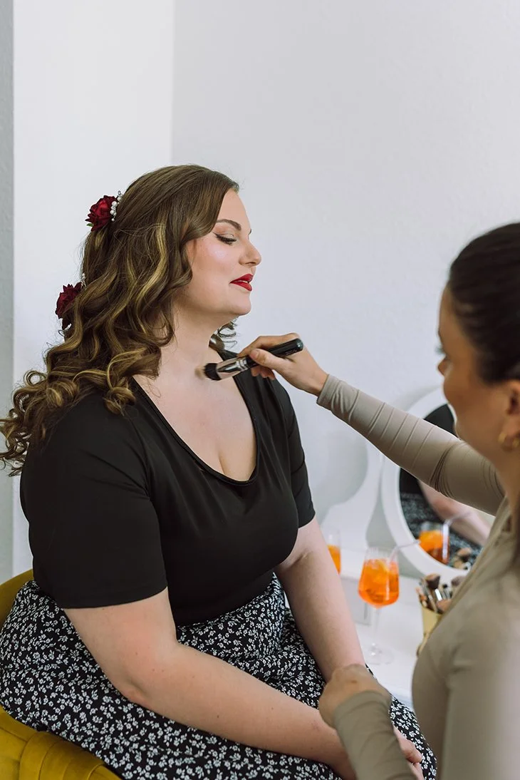 Frau wird im Beauty-Studio von einer Kosmetikerin geschminkt, im Hintergrund ein Spiegel mit Getränken.
