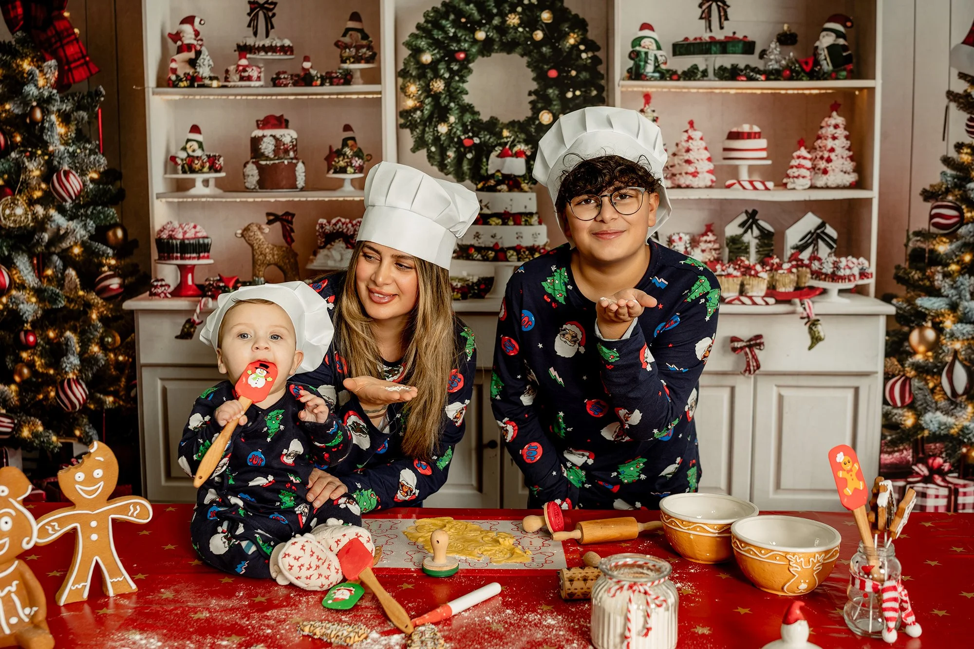 Familie bereitet Weihnachtsplätzchen in festlich geschmückter Küche mit Weihnachtsbaum und Dekoration im Hintergrund. Es tragen Schlafanzüge mit Weihnachtsmotive und Kochhauben.