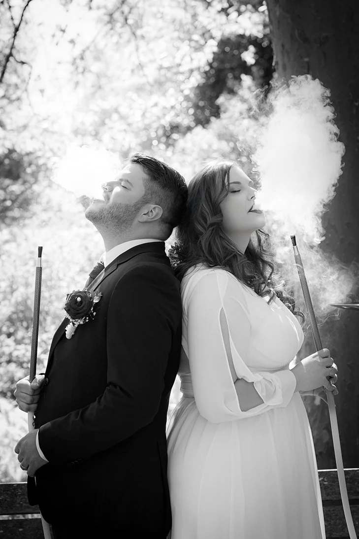 Ein Mann im Anzug und eine Frau im weißen Kleid stehen Rücken an Rücken, beide rauchen, bei Sonnenlicht im Freien, schwarz-weiß Foto, romantische Stimmung.