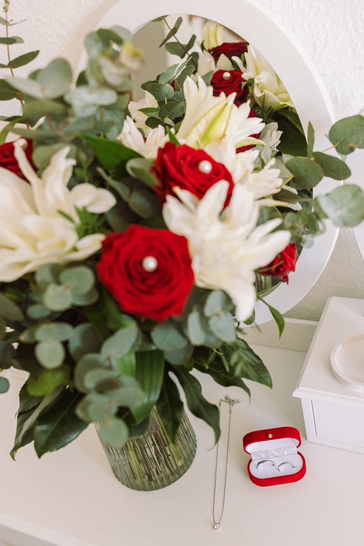 Blumenstrauß aus weißen und roten Blumen mit grünen Blättern vor einem Spiegel, daneben eine offene rote Schmuckschachtel mit zwei Ringen.
