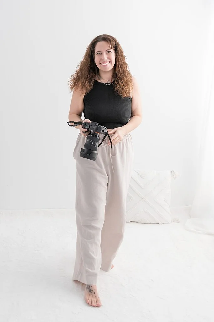 Eine lachende Frau mit lockigem braunem Haar hält eine professionelle Kamera, steht barfuß auf einem weißen Boden vor einem weißen Hintergrund.