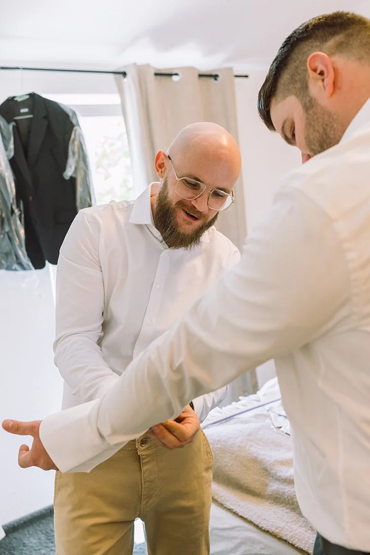 Zwei Männer beim Anziehen eines Hemdes, in einem Zimmer mit Kleidung im Hintergrund.
