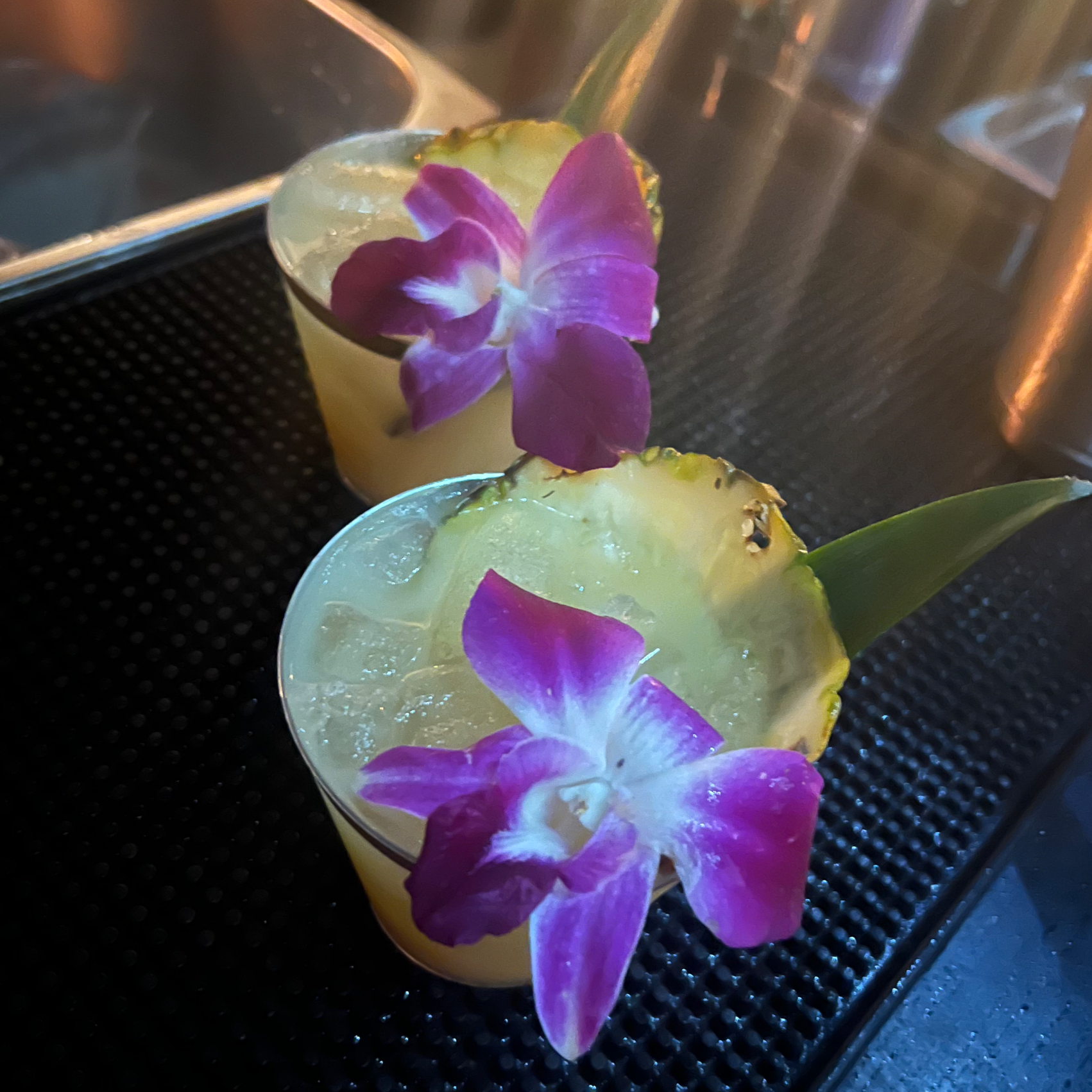 Two glasses filled with a yellowish beverage, garnished with purple orchids and pineapple slices, on a black surface.