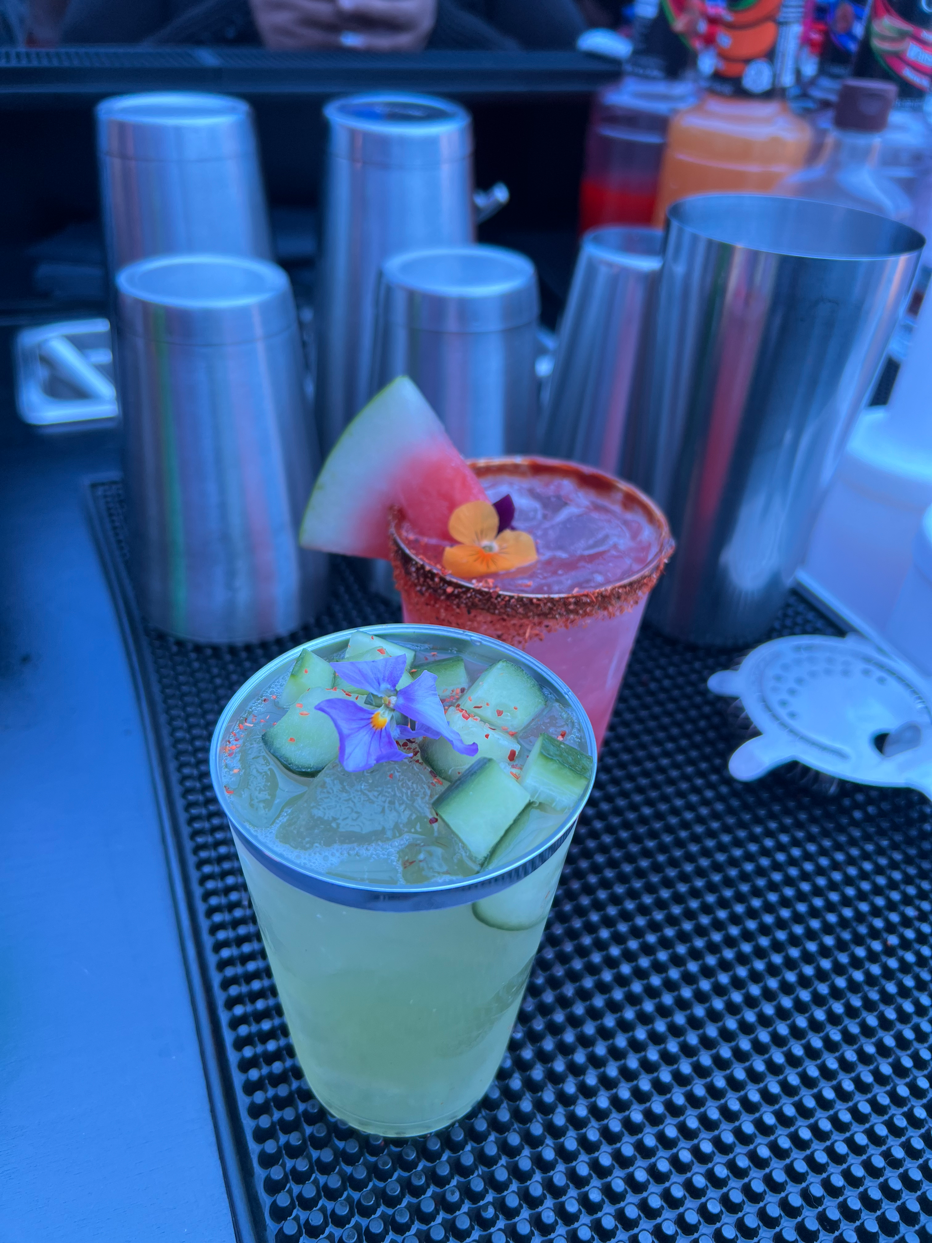 A cocktail garnished with edible flowers, served with cucumber slices in a glass with ice, next to a watermelon slice with an edible flower on top.