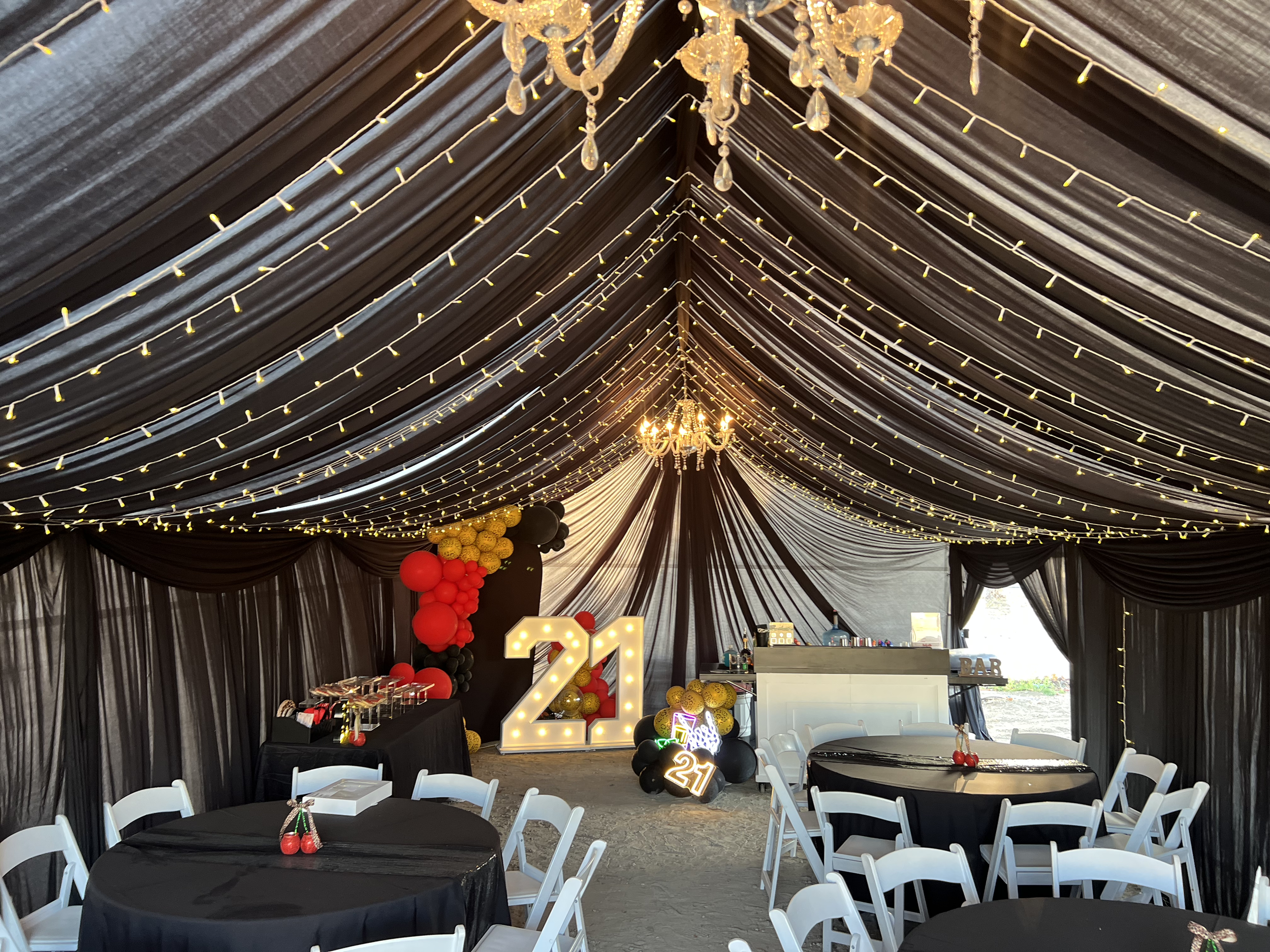 Interior of a decorated event tent with black ceiling drapes and string lights, featuring a large illuminated '21' sign and balloon arrangements in black, red, and gold, set up for a celebration or party.