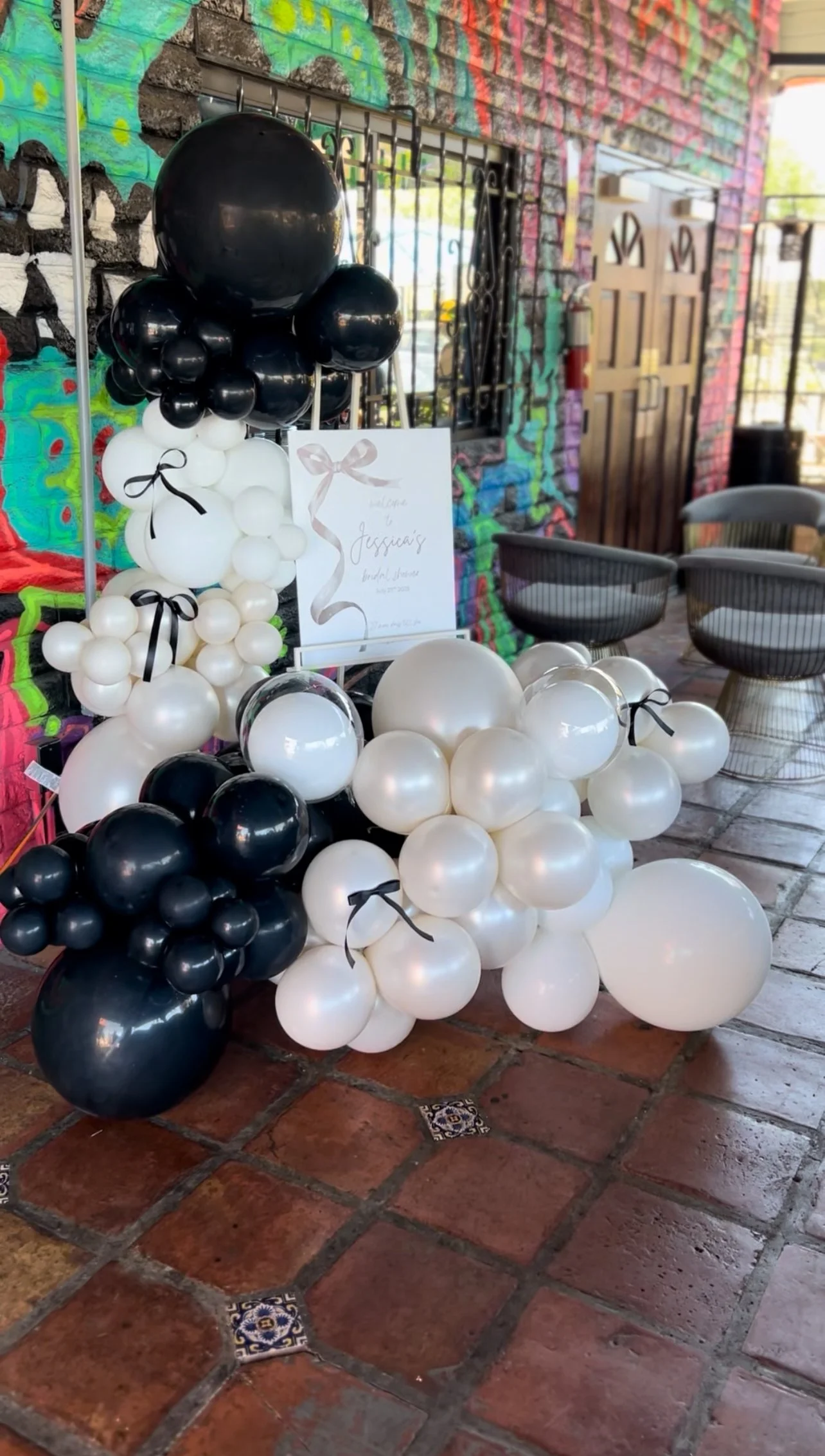 A balloon decoration arrangement with black, white, and silver balloons, some tied with black ribbons, set against a colorful graffiti wall and brick floor. There is a sign with cursive writing and a pink ribbon design, indicating a celebration.