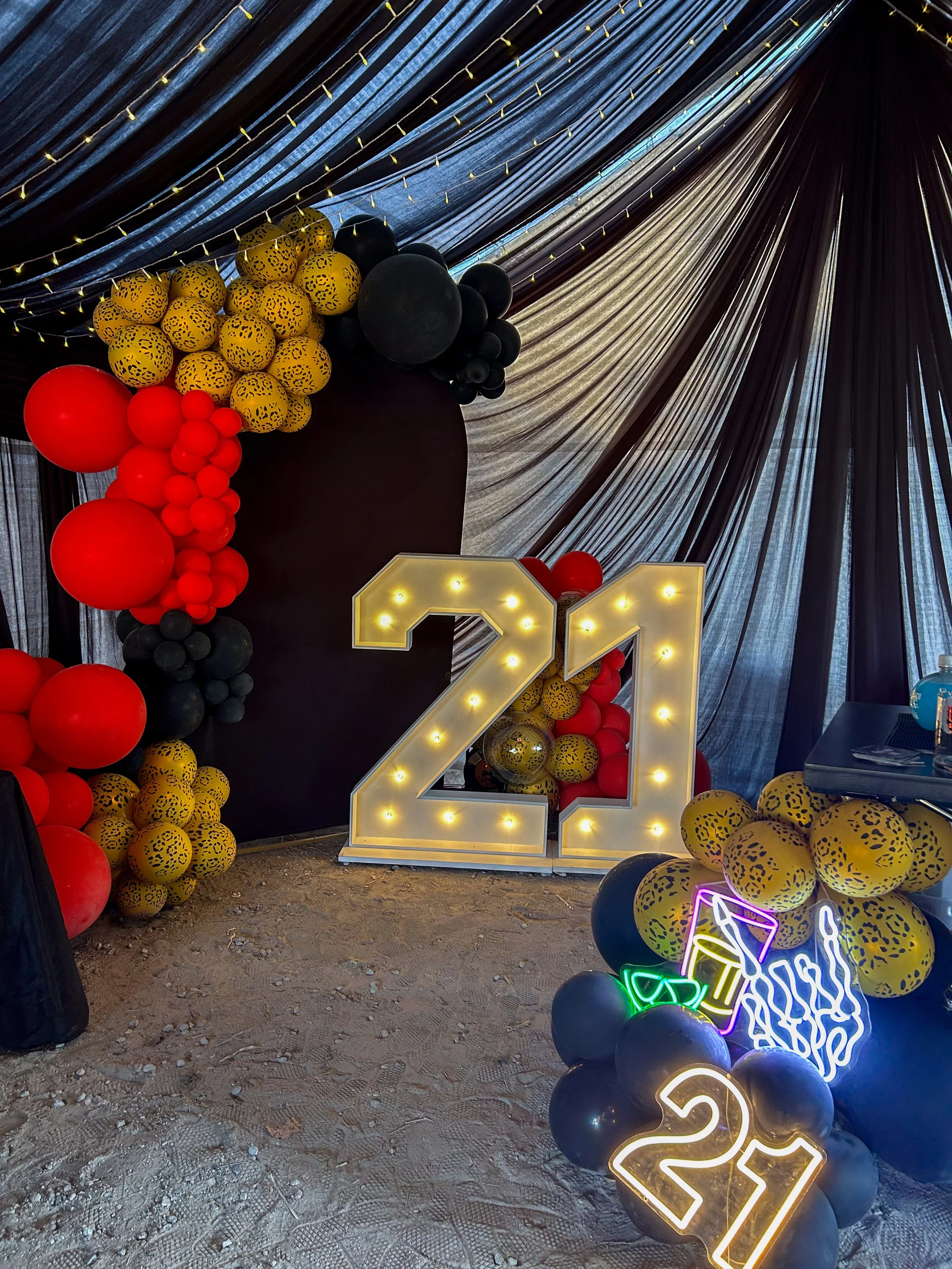 Decorative celebration setup with a large illuminated number 21, surrounded by balloons in red, black, and yellow with black leopard print patterns, and neon signs with drinks and party symbols, under black draped fabric ceiling.