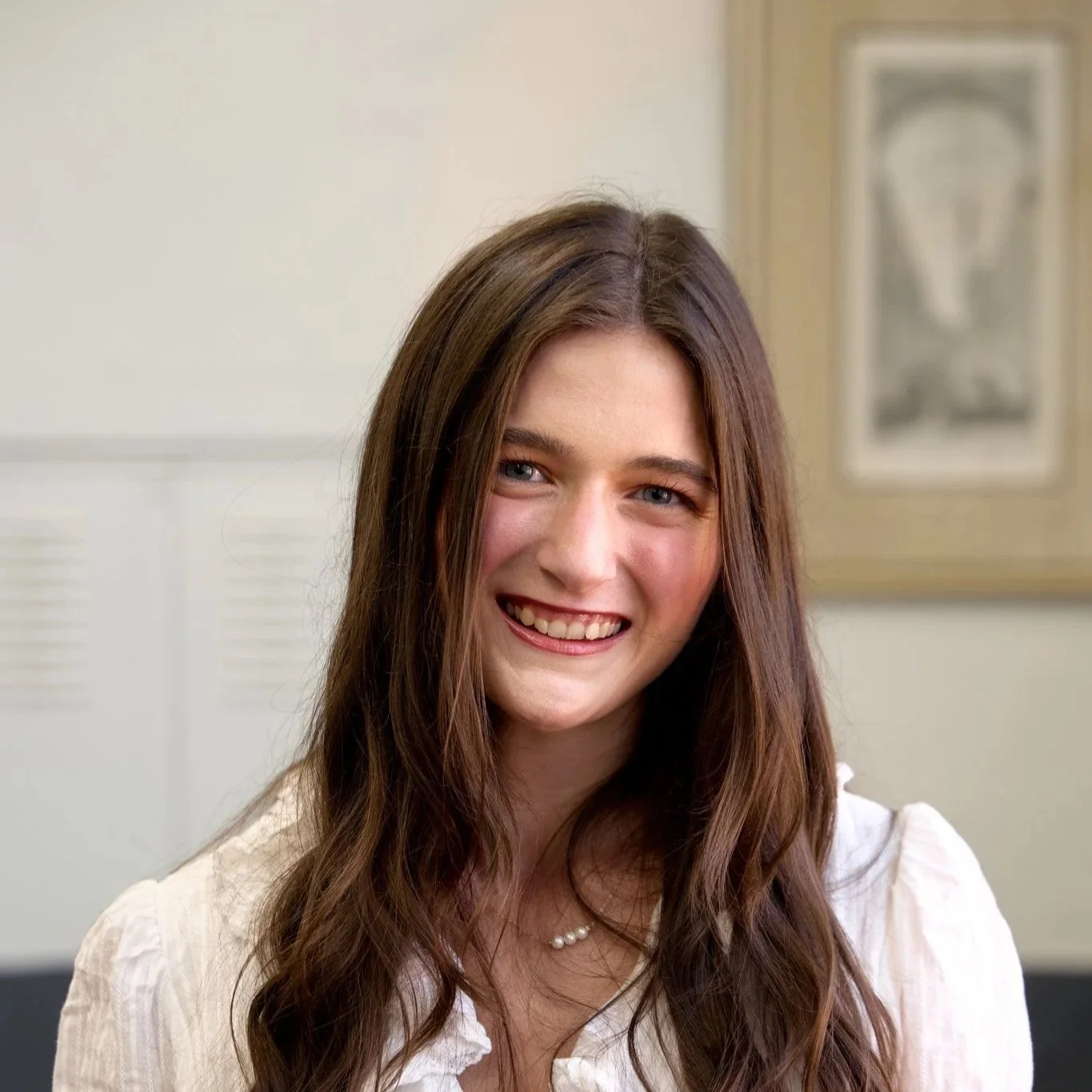 A young woman with long wavy brown hair smiling, wearing a black top and a pearl necklace, standing indoors.