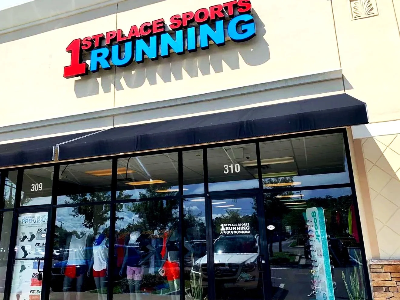 Exterior of a retail store named 1st Place Sports Running with large red and blue lettering on the sign, glass storefront, and mannequins inside displaying athletic apparel.