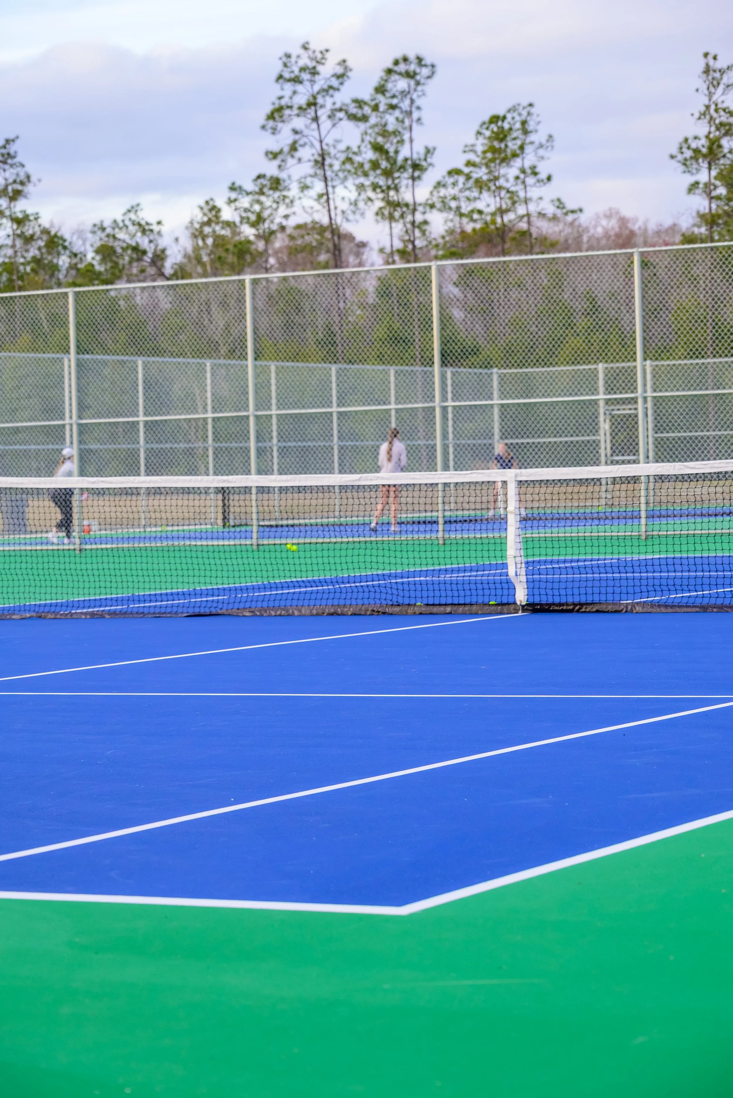 Colorful outdoor tennis court with blue and green surface, chain-link fencing, and people practicing tennis in the background, with trees and cloudy sky.