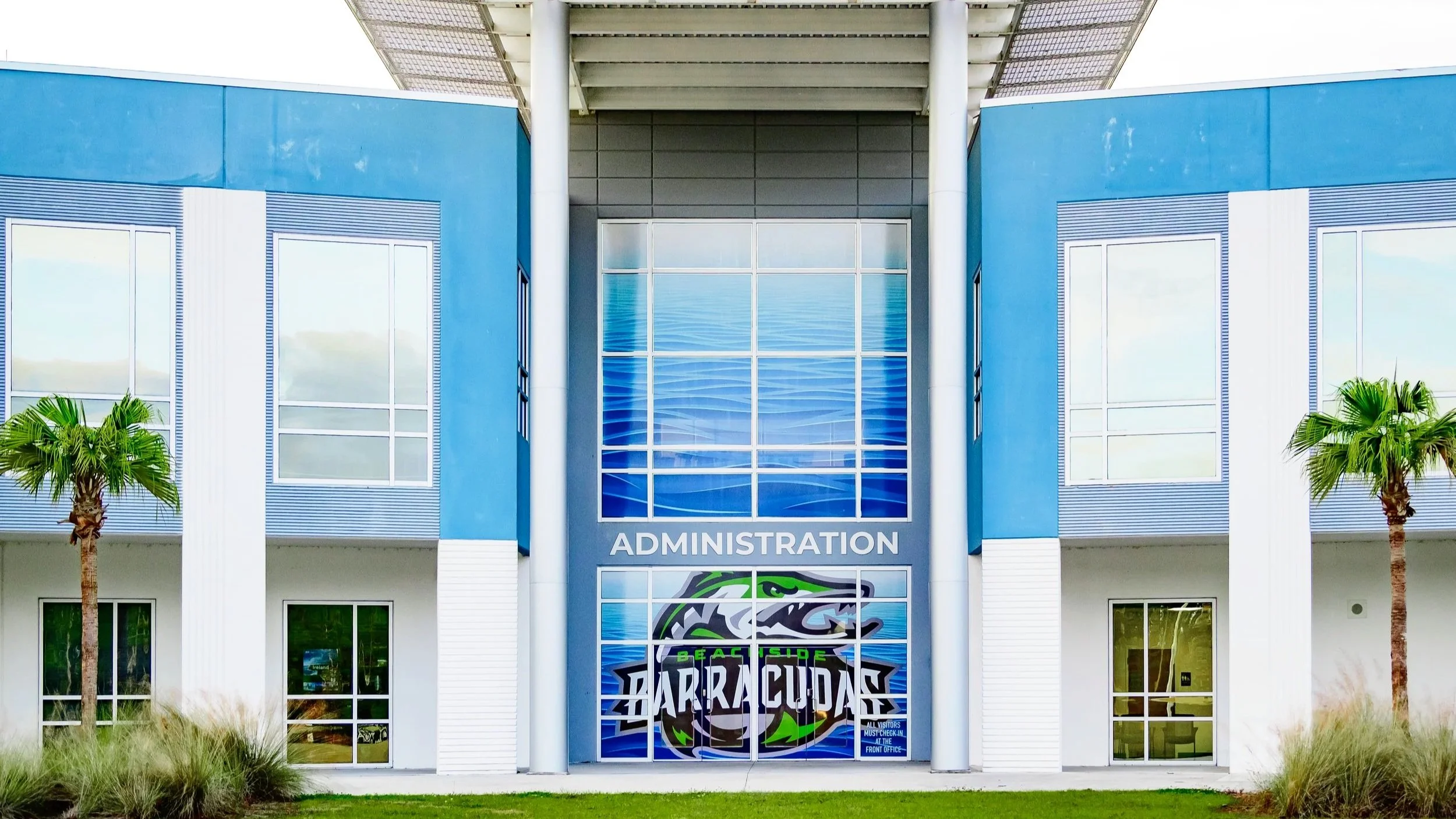 The building of  Beachside HS features a large blue facade with windows and palm trees on each side. The entrance has a mural of a stylized animal head with the words 'Beachside Barracudas' and a sign indicating 'Administration' above the door.