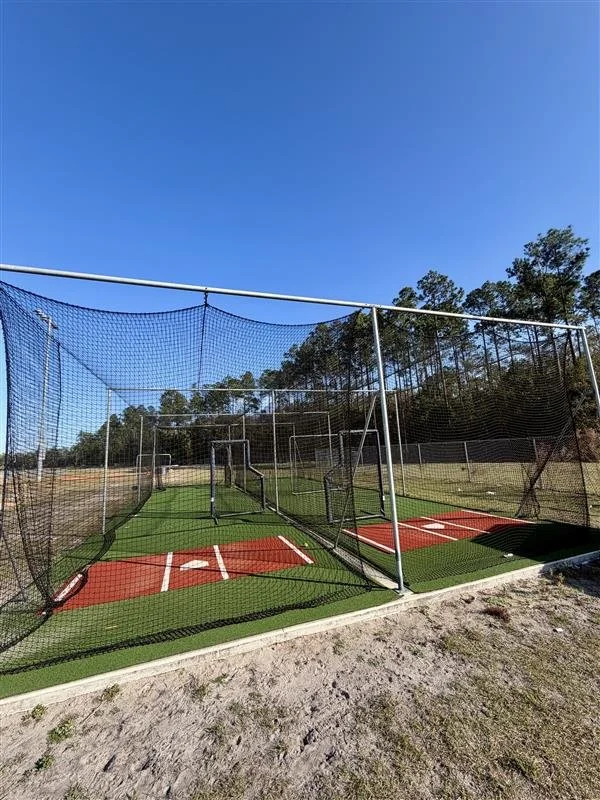 A baseball practice cage with two batting areas on artificial turf, enclosed by mesh netting and surrounded by trees and open field under a clear blue sky.