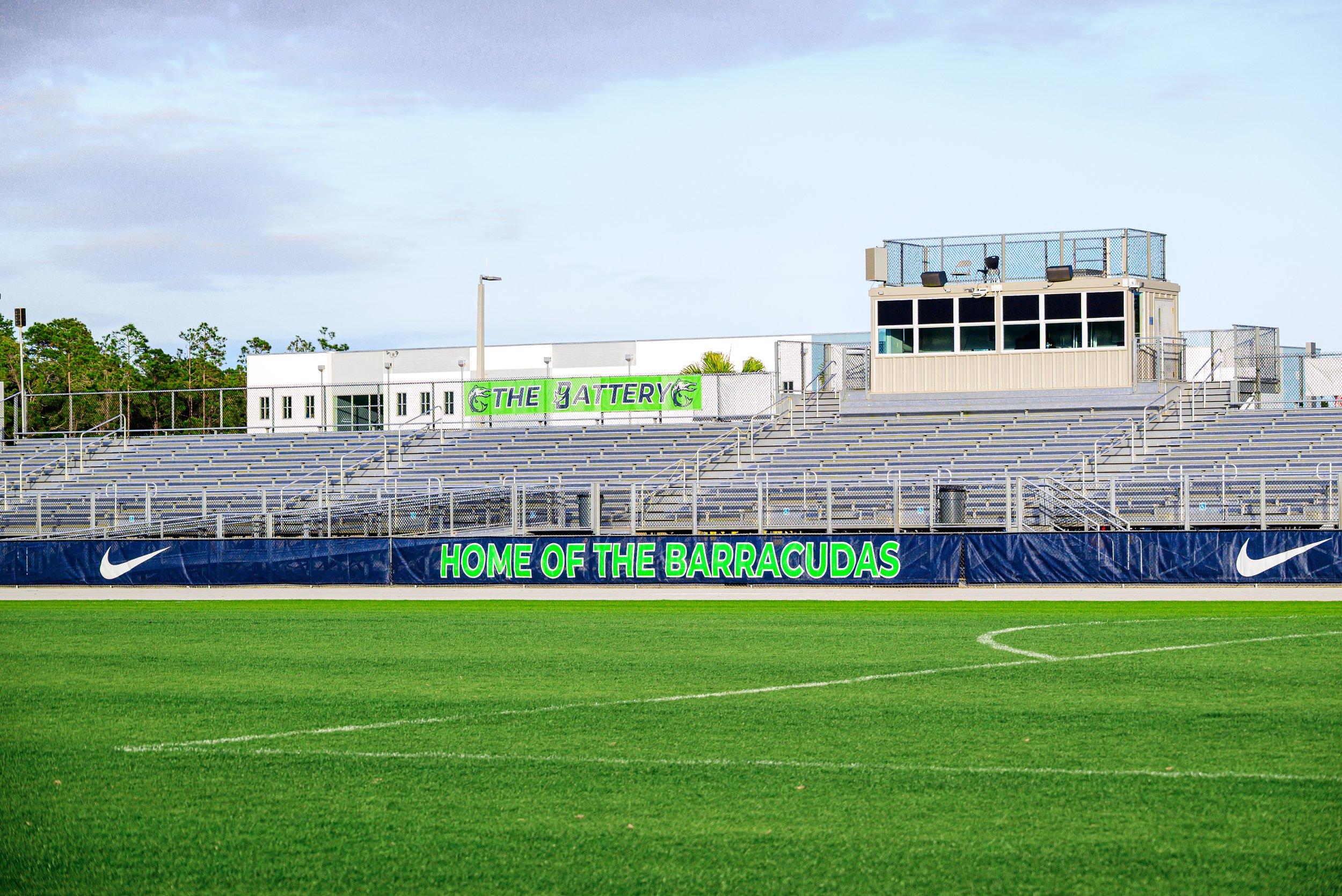Empty sports stadium bleachers with a sign that reads 'Home of the Barracudas' and a building labeled 'The Battery' in the background, with a green grassy field in the foreground.