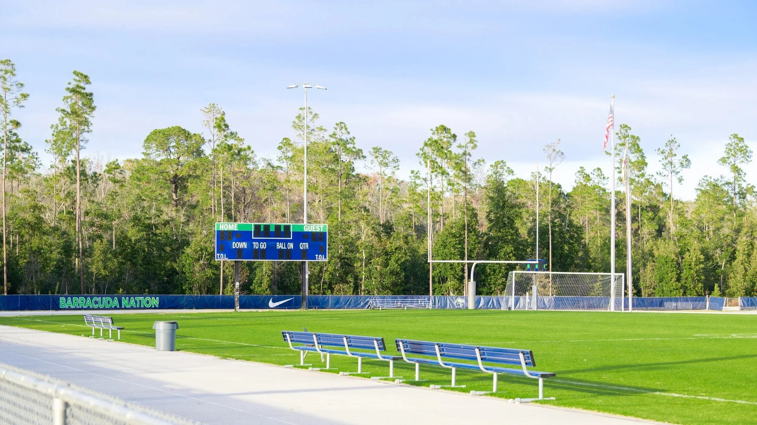 Empty outdoor sports field with green grass, benches, goalpost, and scoreboard, surrounded by trees and blue sky.