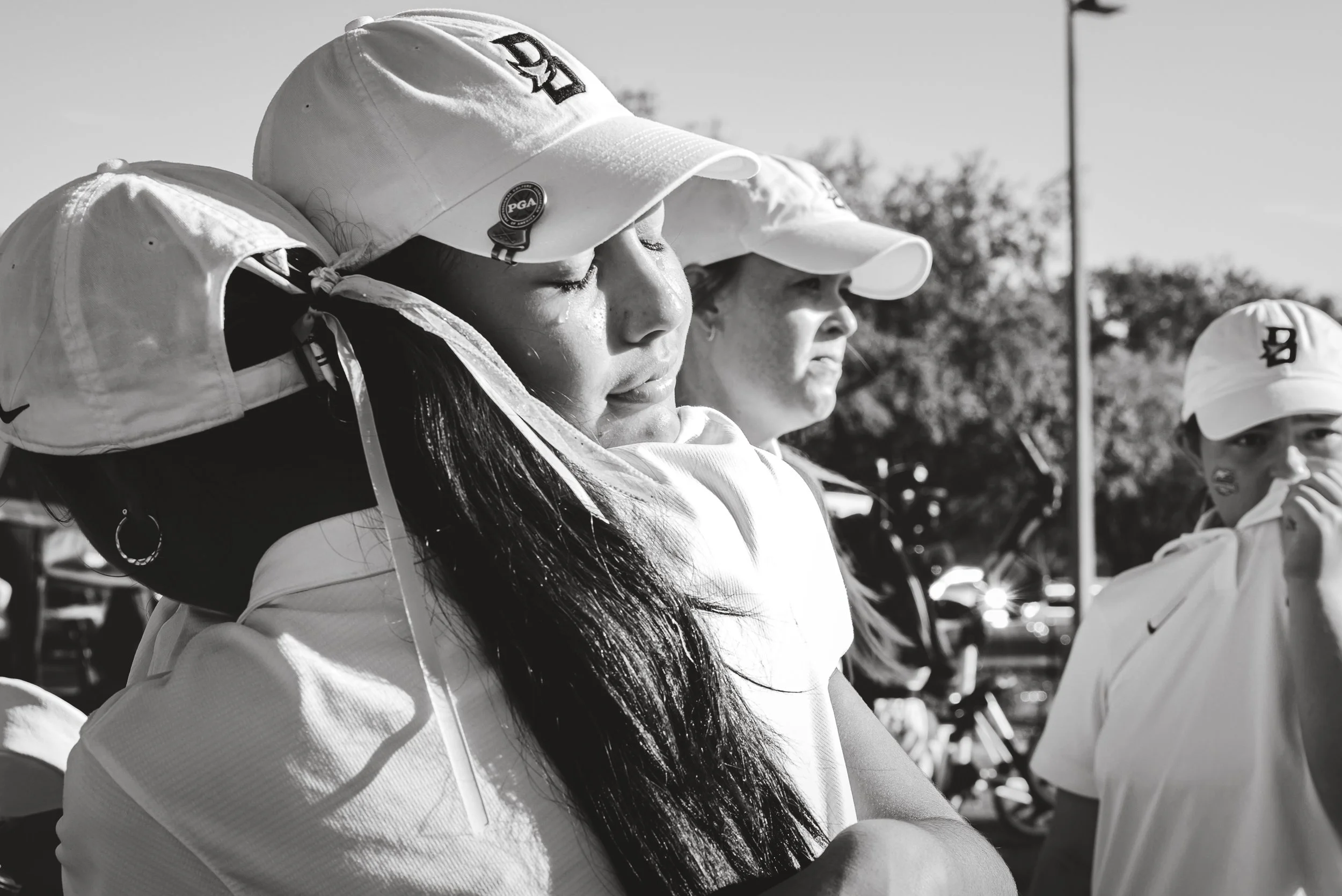 Three young female athletes wearing caps and sports attire look downcast with somber expressions. They are outside, with trees and parked cars visible in the background.
