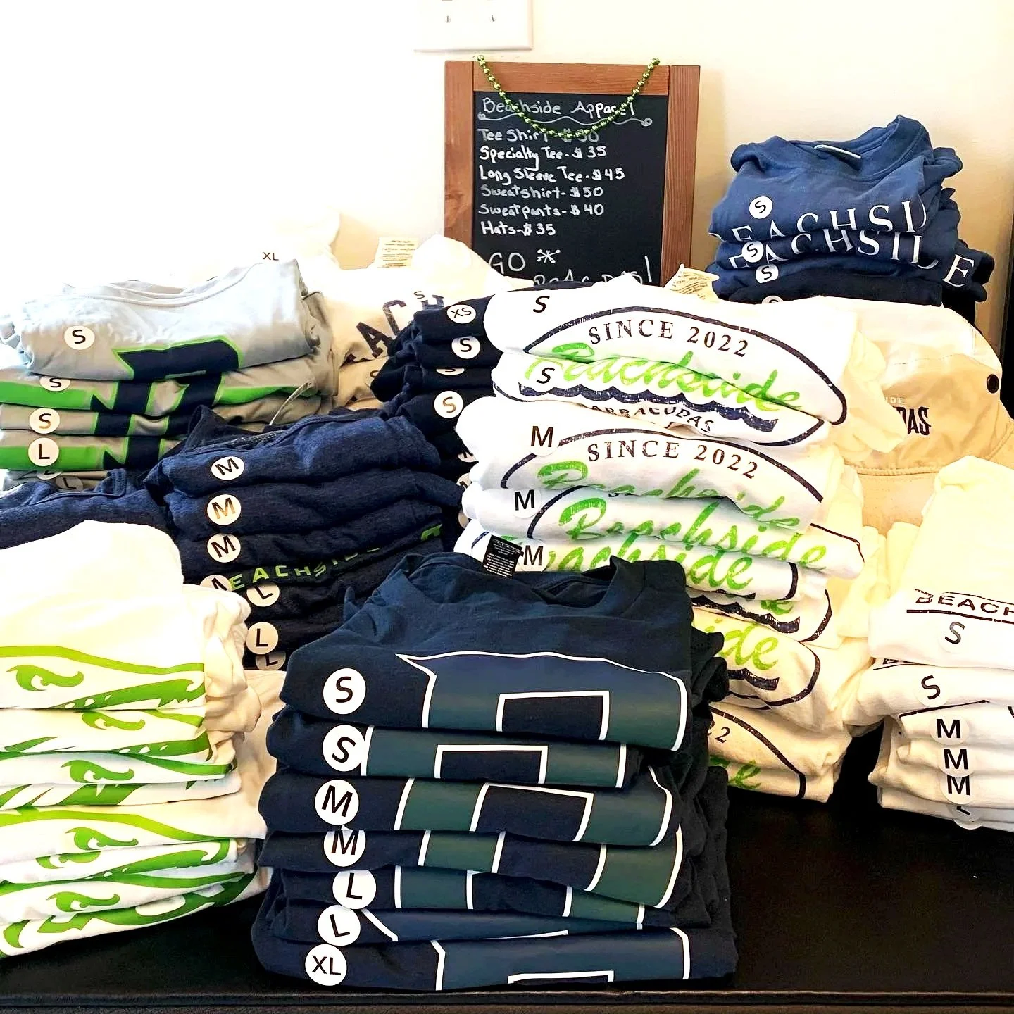 Stacks of folded t-shirts in various colors including white, navy blue, gray, and black, with some displaying the word 'Beachside' and others showing stylized designs. A small chalkboard sign in the background lists prices and items available for sale.