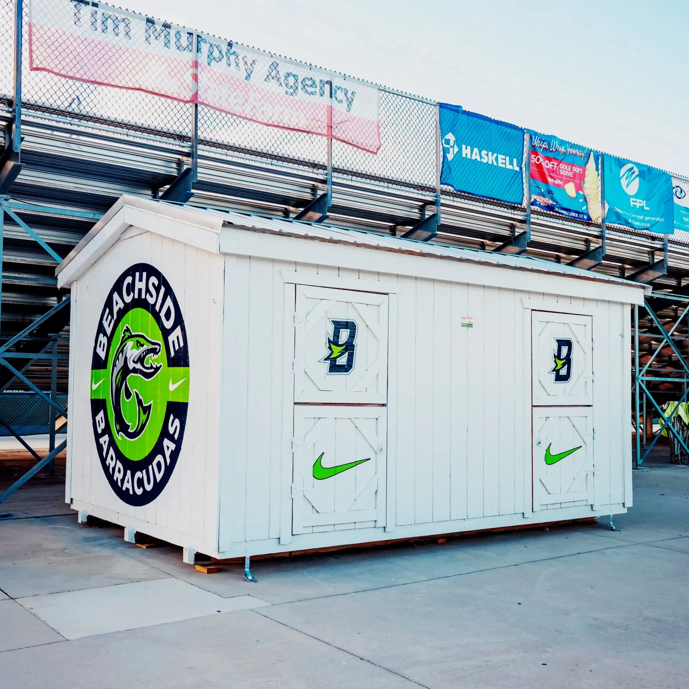 white structure with logos and designs, likely a stadium or event area, with a large circular Beauchide Barracudas logo on the side, featuring a stylized barracuda fish head in green and black. The front panels have logos including a blue and white emblem with a star and leaf, and a green Nike swoosh. In the background, there are advertisements and a section of stadium seating.