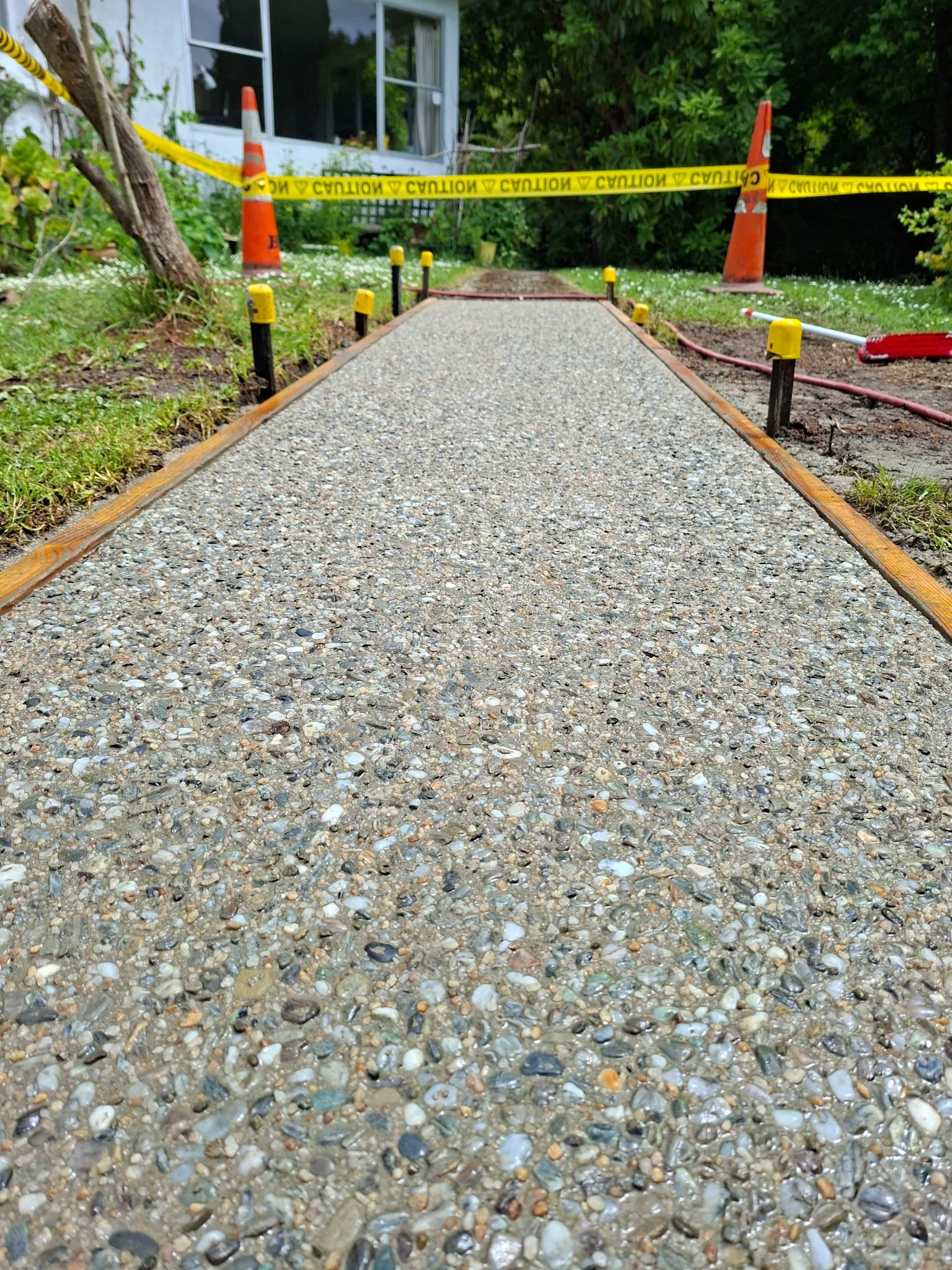 A newly poured concrete sidewalk in front of a house, with orange traffic cones, caution tape, and construction equipment around it, indicating ongoing construction work.