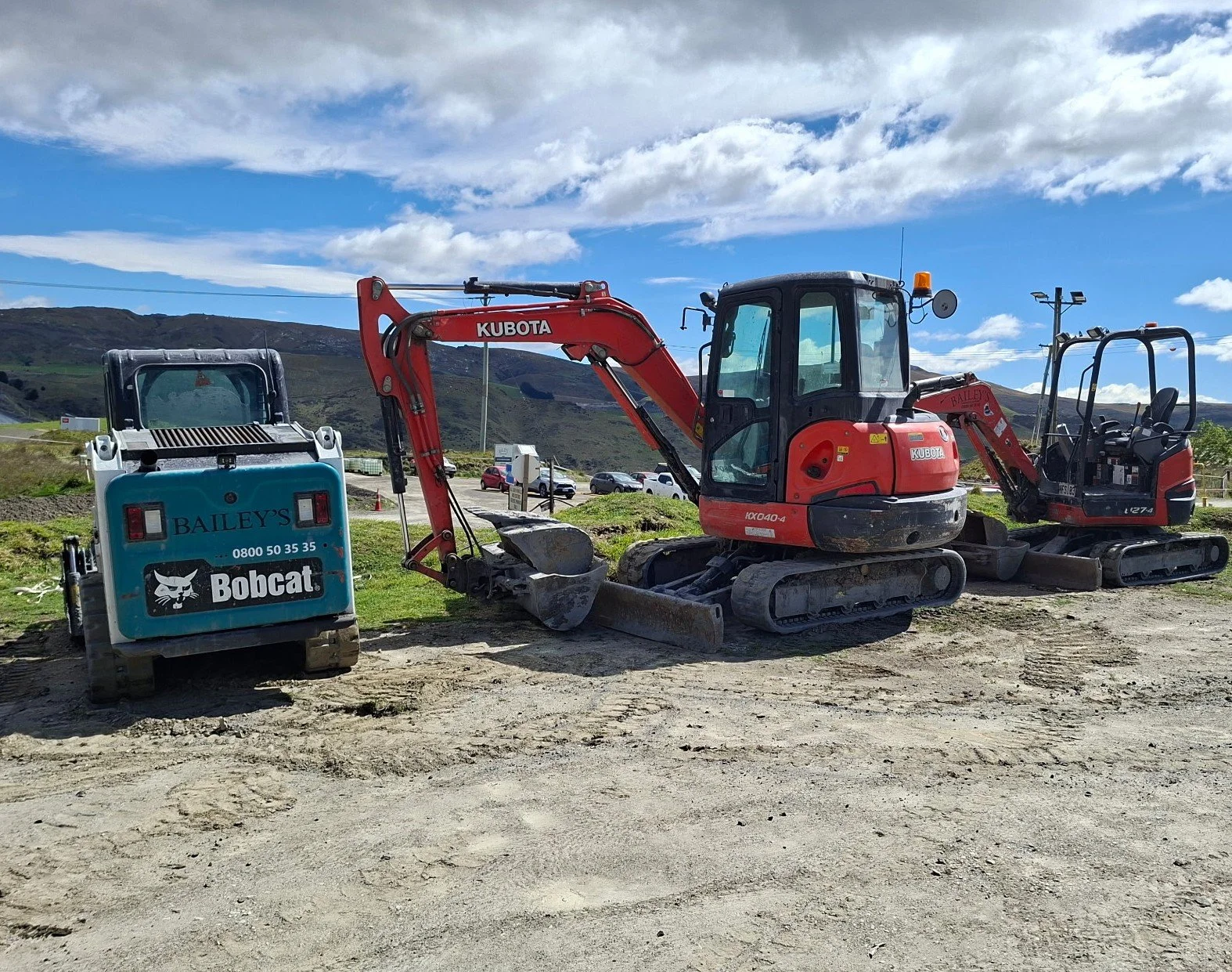 Two small construction excavators, one with a red arm and the other with a black arm, are parked on a dirt lot with a mountainous landscape and cloudy sky in the background.