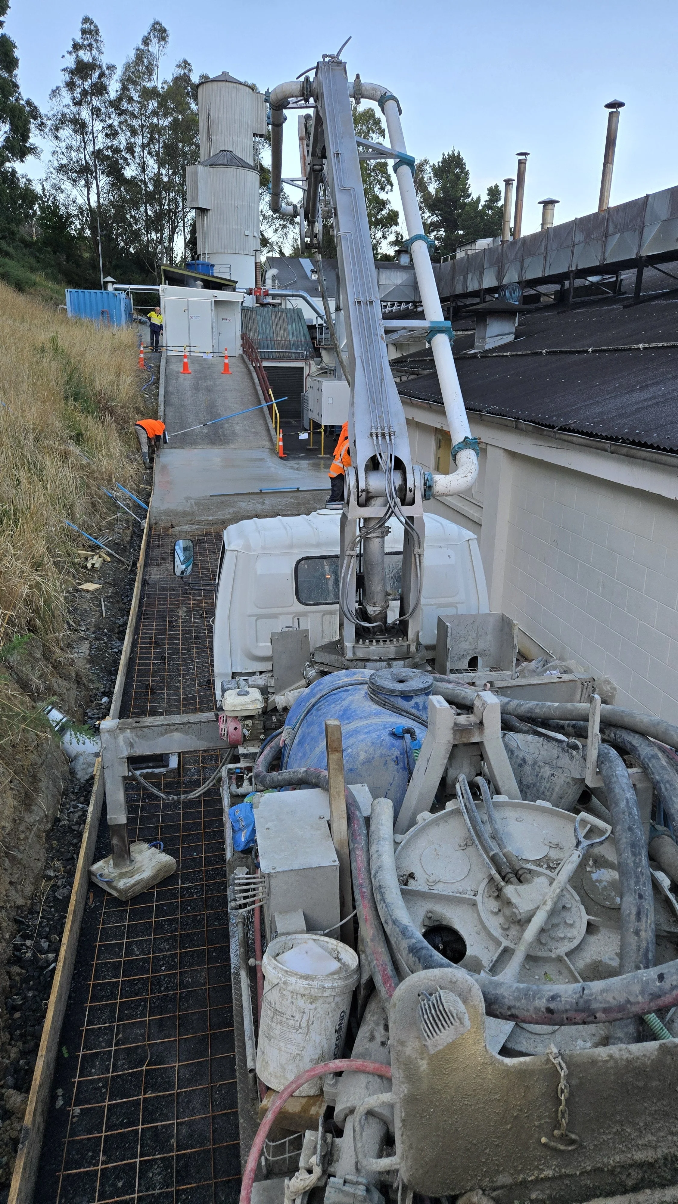 Construction site with workers paving a walkway, machinery, and piping, alongside a building and hillside with trees.