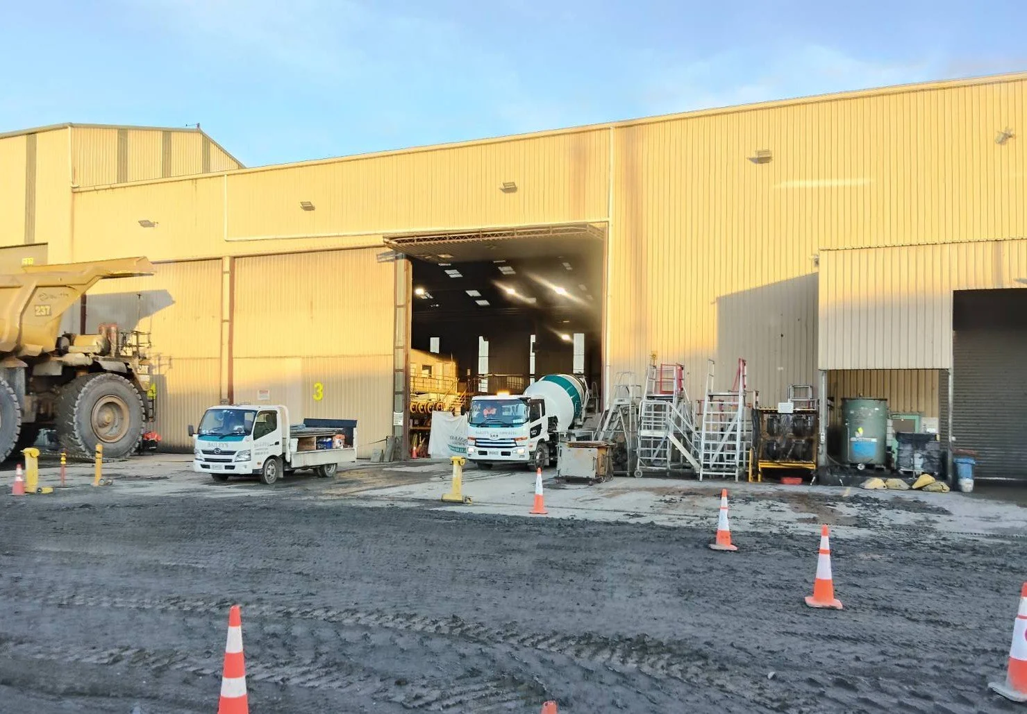 Construction site with a yellow industrial building, construction vehicles, trucks, and various construction equipment, with traffic cones and a muddy foreground.