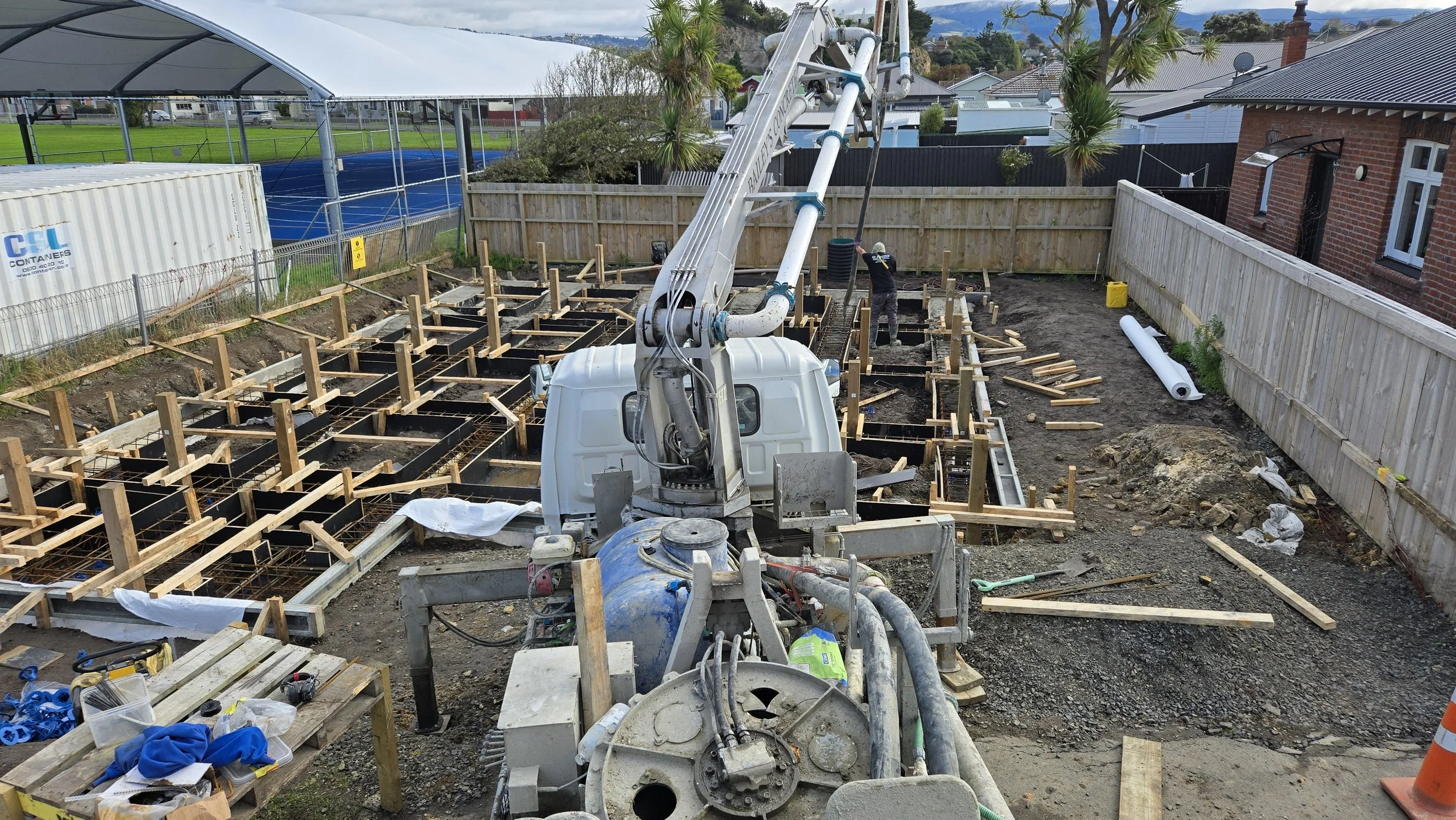 Construction site with a concrete pouring pump, wooden formwork, and an excavated foundation area surrounded by a wooden fence, with houses and trees in the background.
