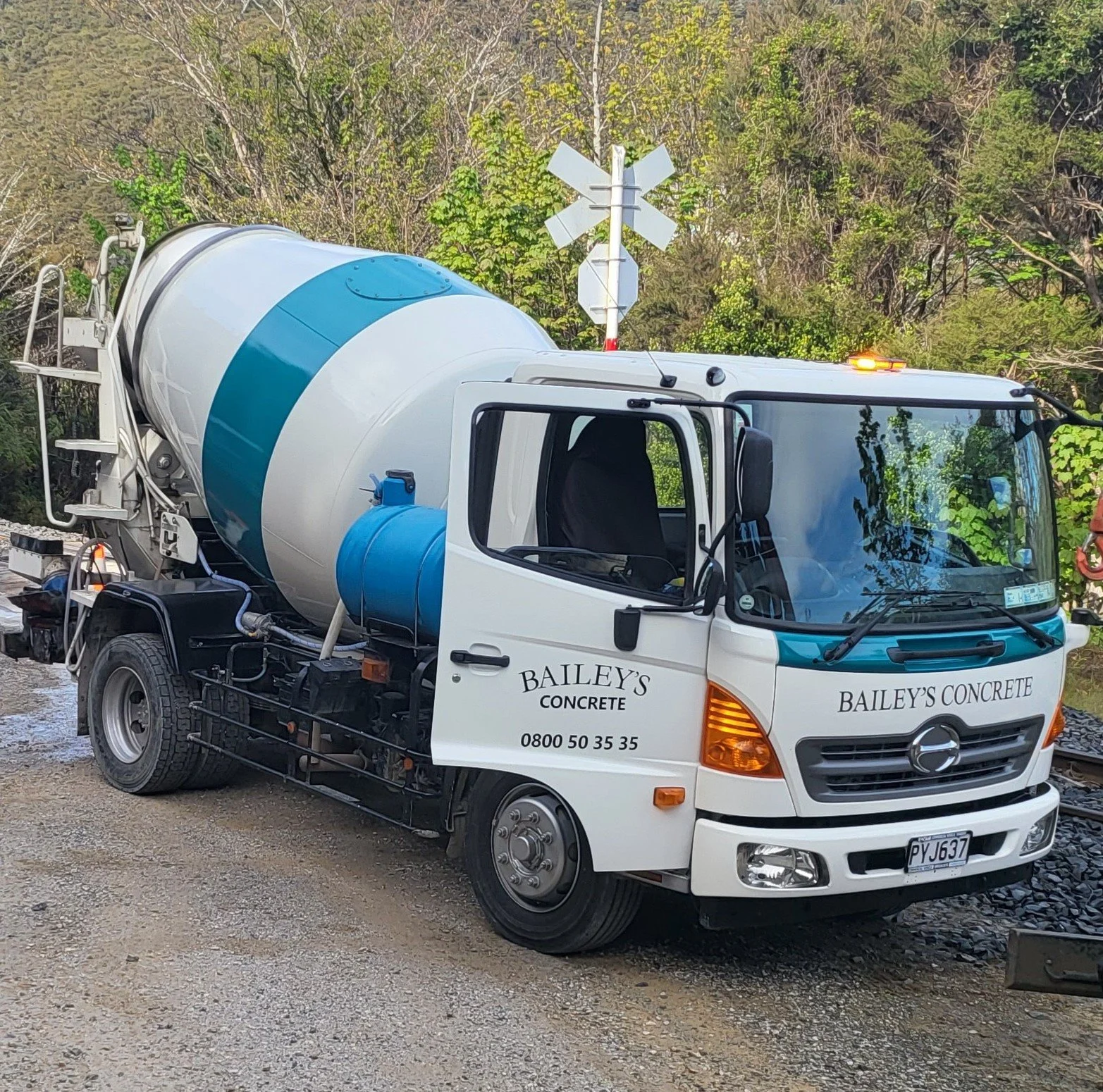A white cement mixer truck from Bailey's Concrete parked on a gravel surface with a railroad crossing sign and trees in the background.