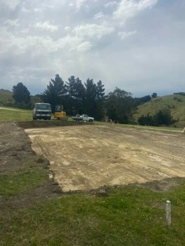 Empty dirt lot with four parked vehicles in the background, trees, hills, partly cloudy sky.
