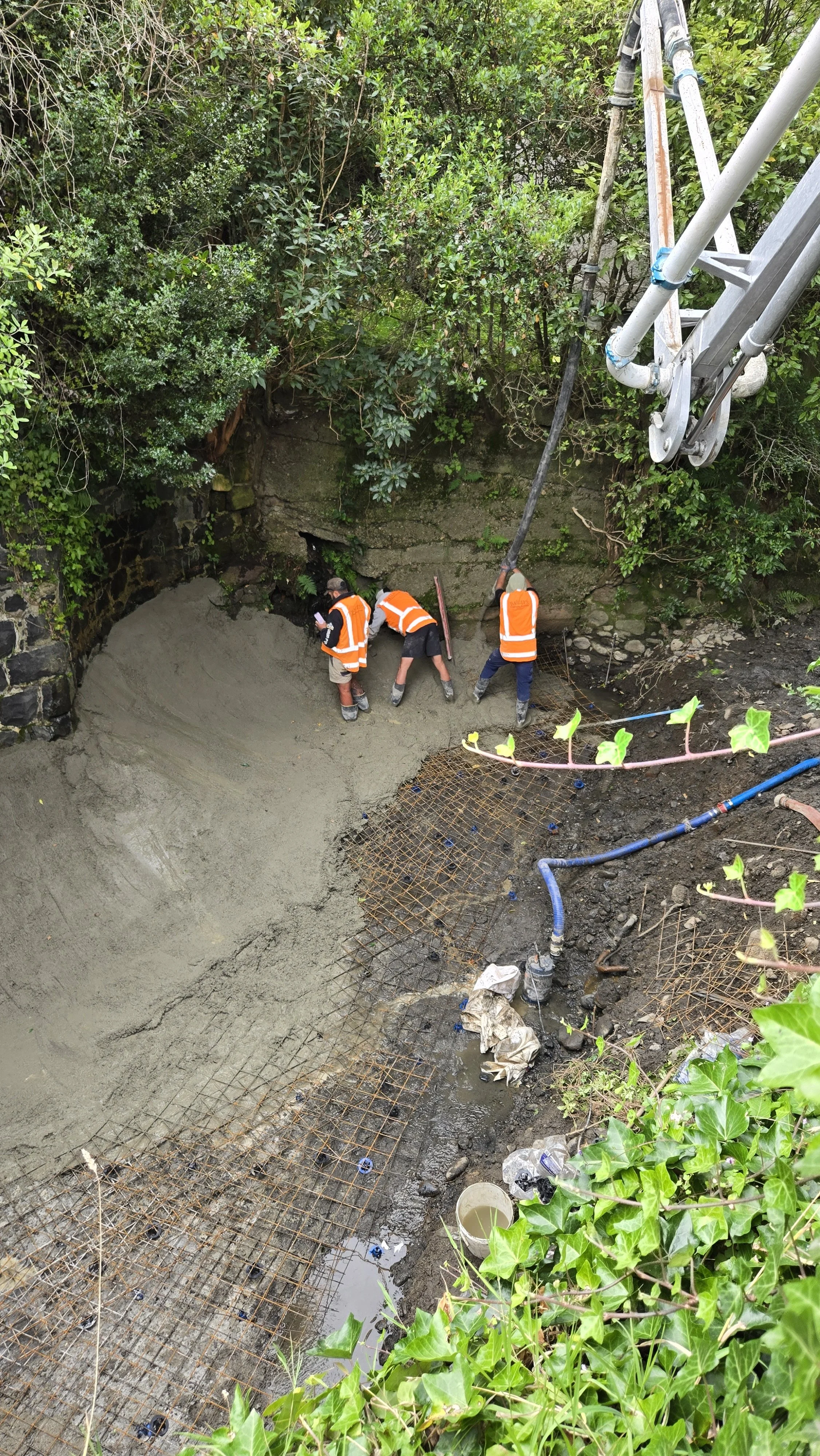 Construction workers with orange safety vests and helmets working on a concrete foundation or pour site, surrounded by greenery, with a large piece of construction equipment overhead and rebar grid in the foreground.
