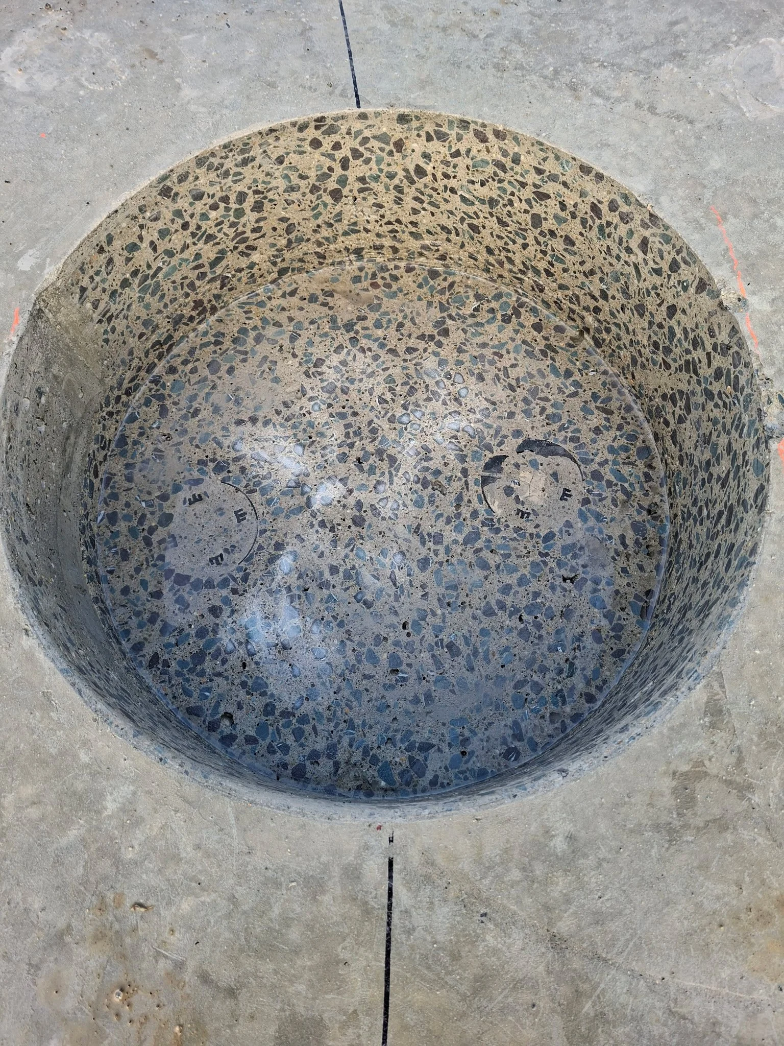 Circular concrete structure with a surface covered in small, multicolored pebbles, embedded in a concrete sidewalk.