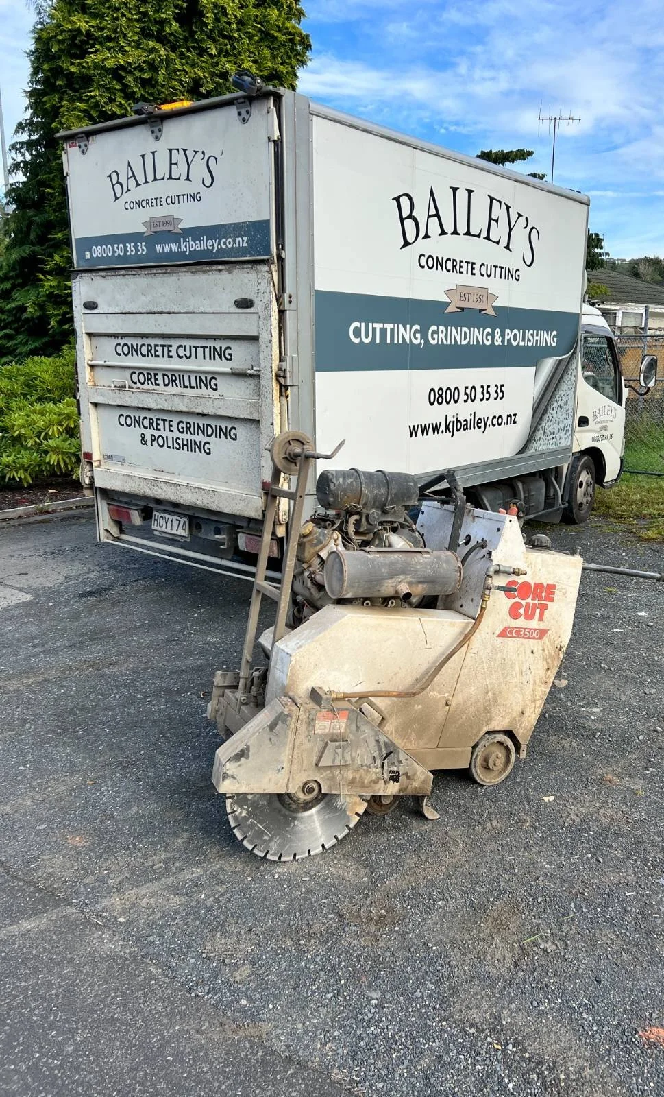 A concrete cutting truck with branding for Bailey's Concrete Cutting parked on a gravel surface beside a small industrial saw machine labeled 'Core Cut' in an outdoor setting with trees and a building in the background.