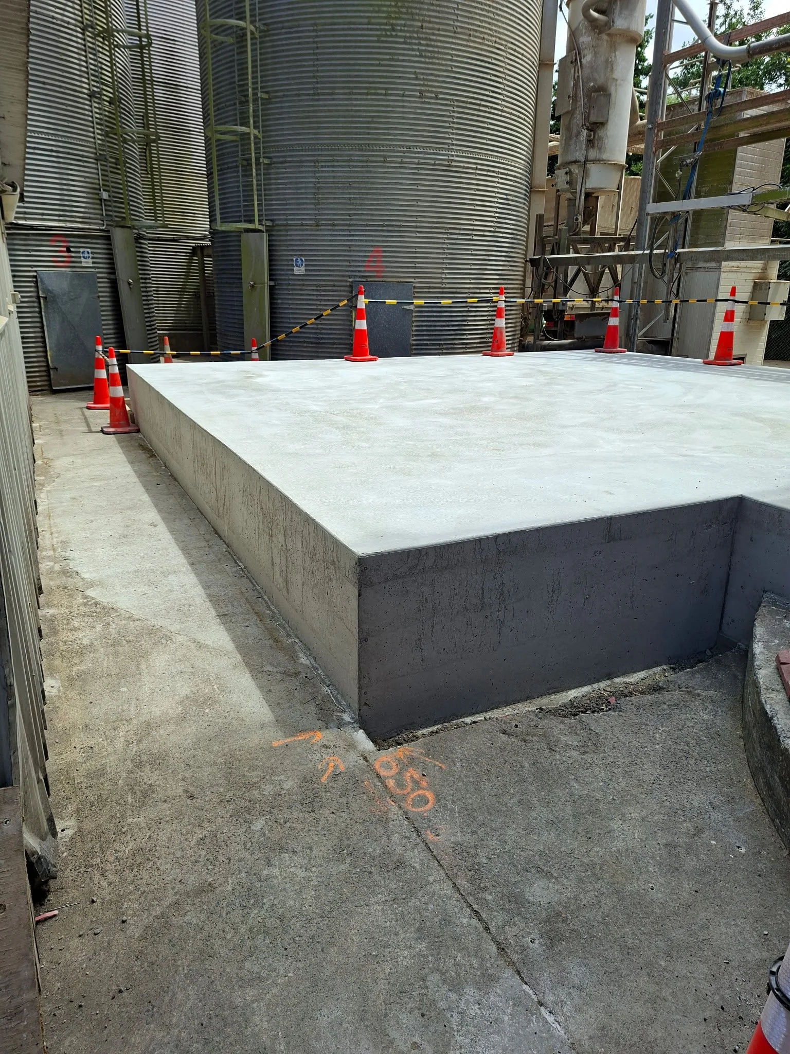 Concrete platform surrounded by orange construction cones and caution tape at an outdoor construction site with large metal storage tanks in the background.