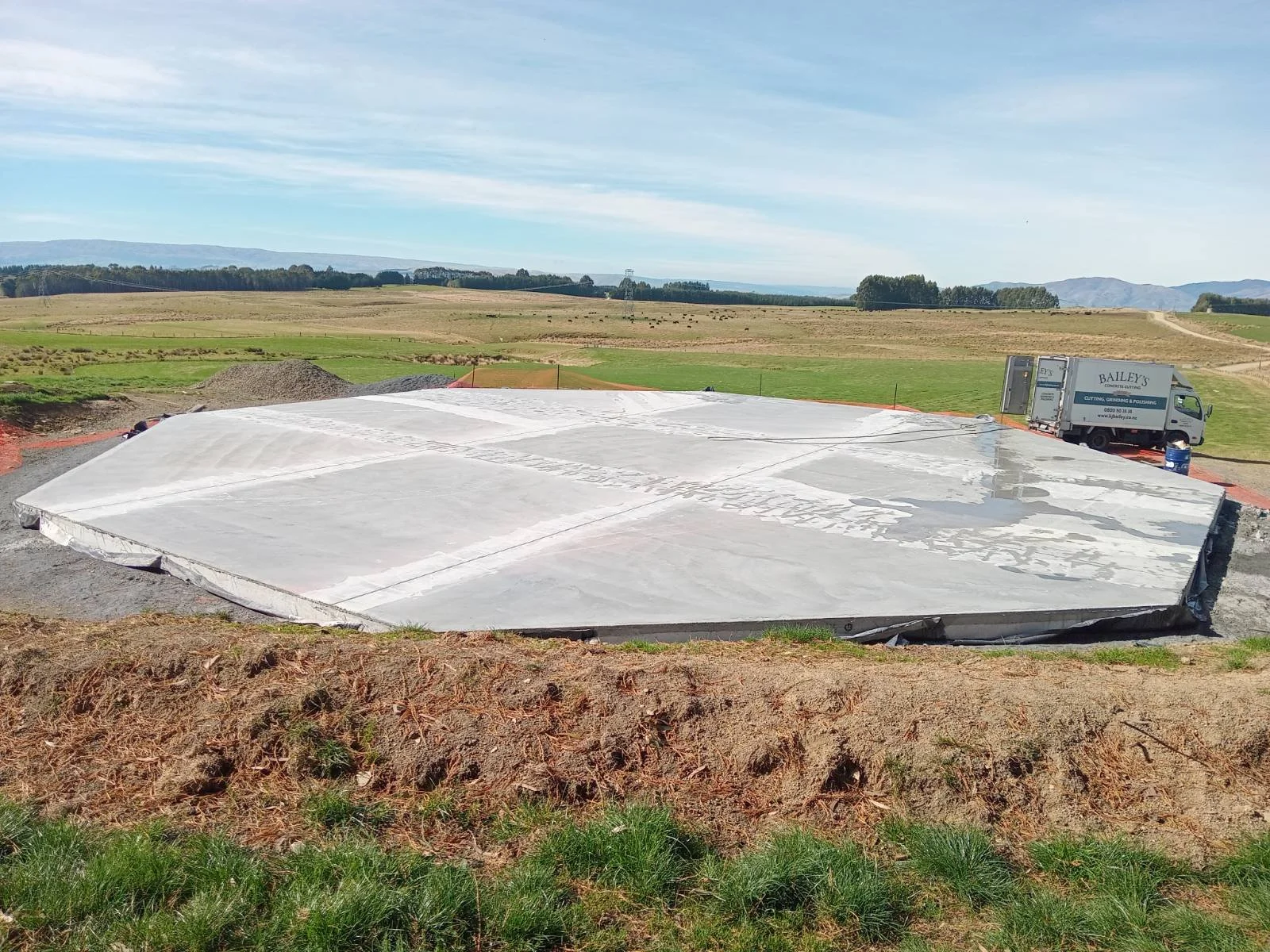 Construction site with a large concrete slab, a Bailey's construction truck, and a rural landscape with open fields and distant hills under a partly cloudy sky.