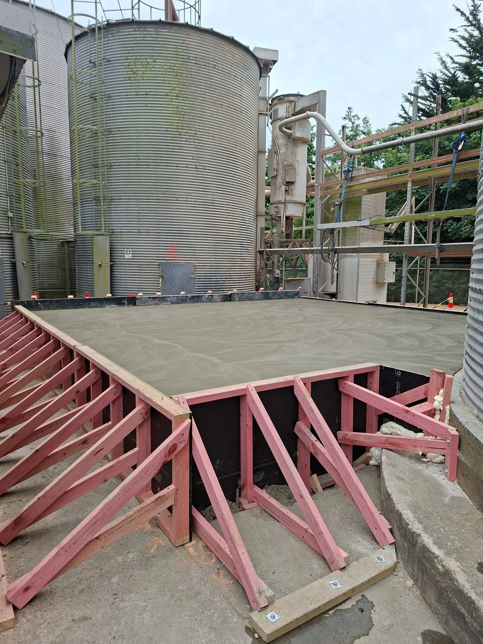Construction site with poured concrete slab, pink wooden forms, and scaffolding around large metal tanks.