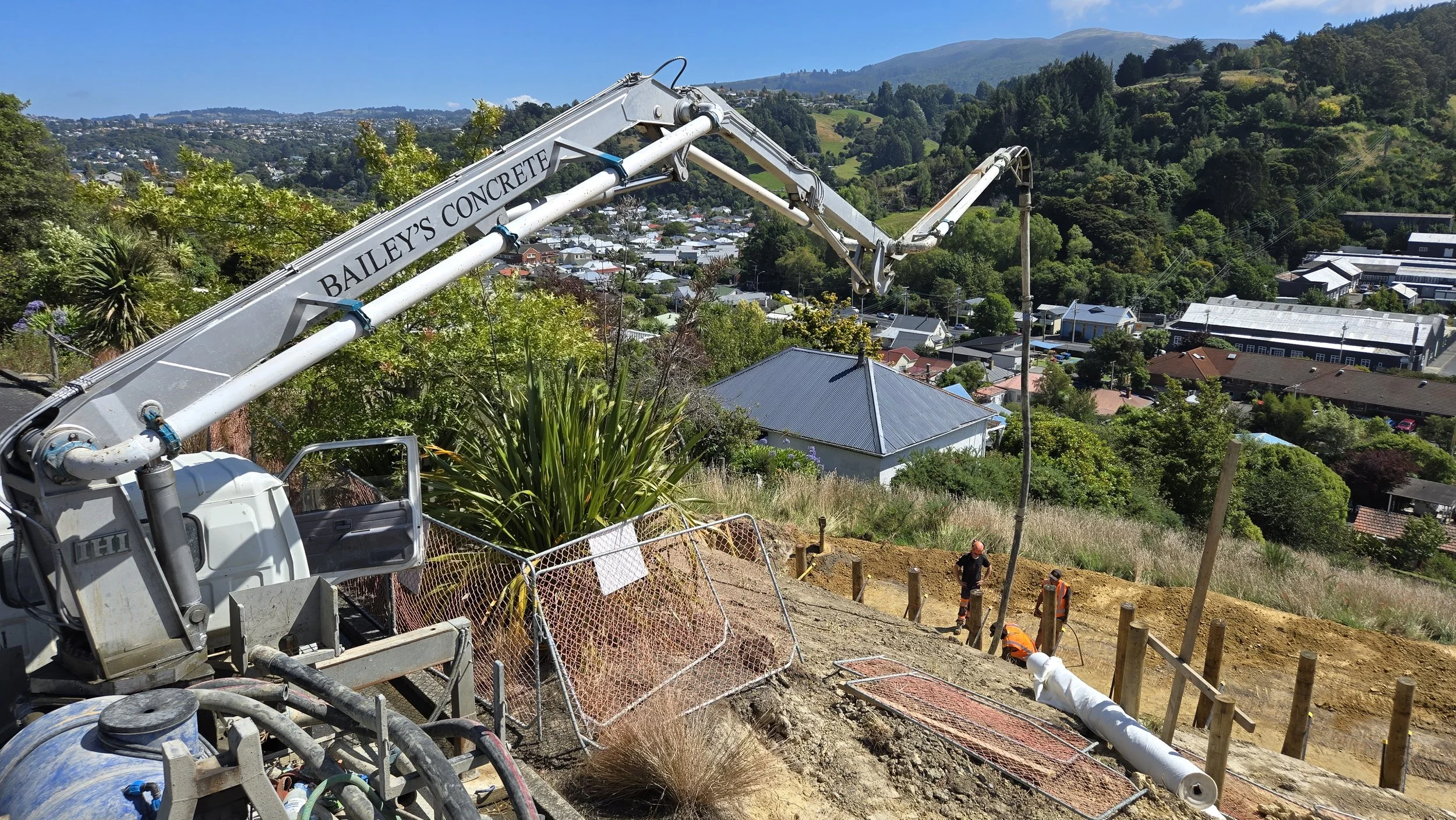 Construction workers and a concrete pump on a hillside construction site overlooking a town with houses, trees, and mountains in the background.