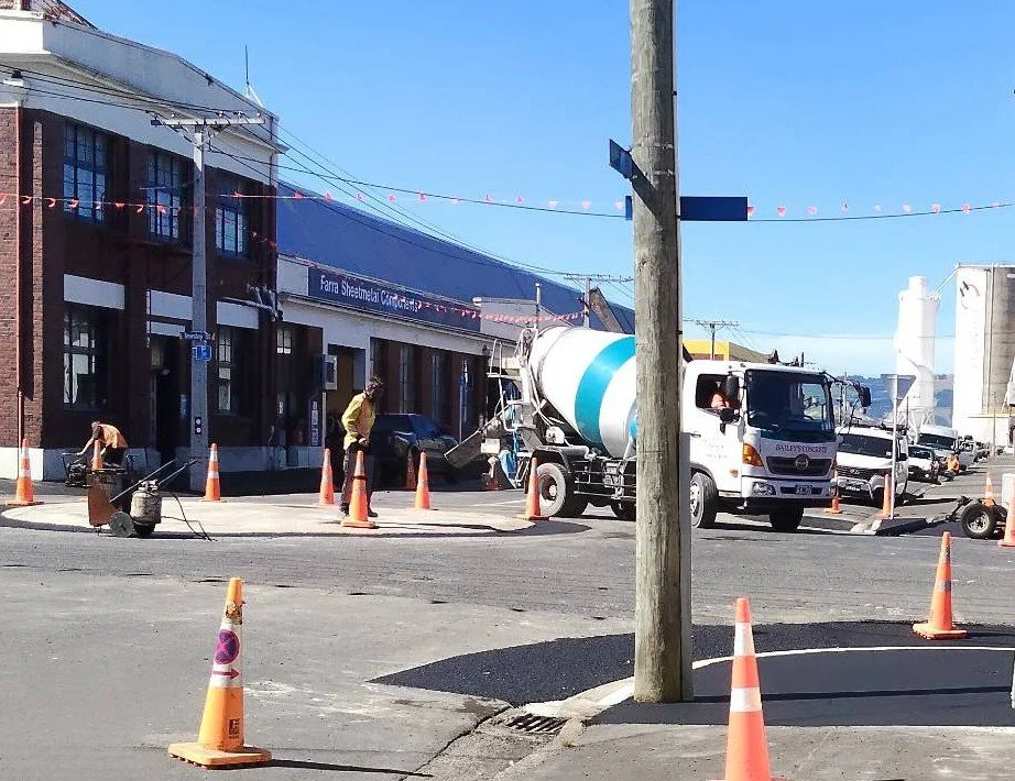 Street construction scene with a concrete mixer truck, construction workers, orange traffic cones, and a building with a sign