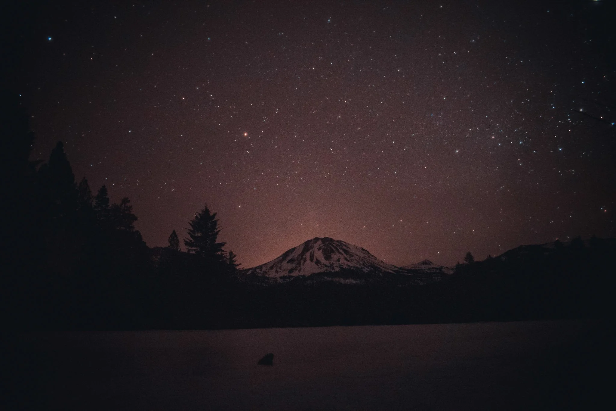 Nighttime landscape with starry sky, silhouette of trees, snow-capped mountain, and a small boat on a calm body of water.