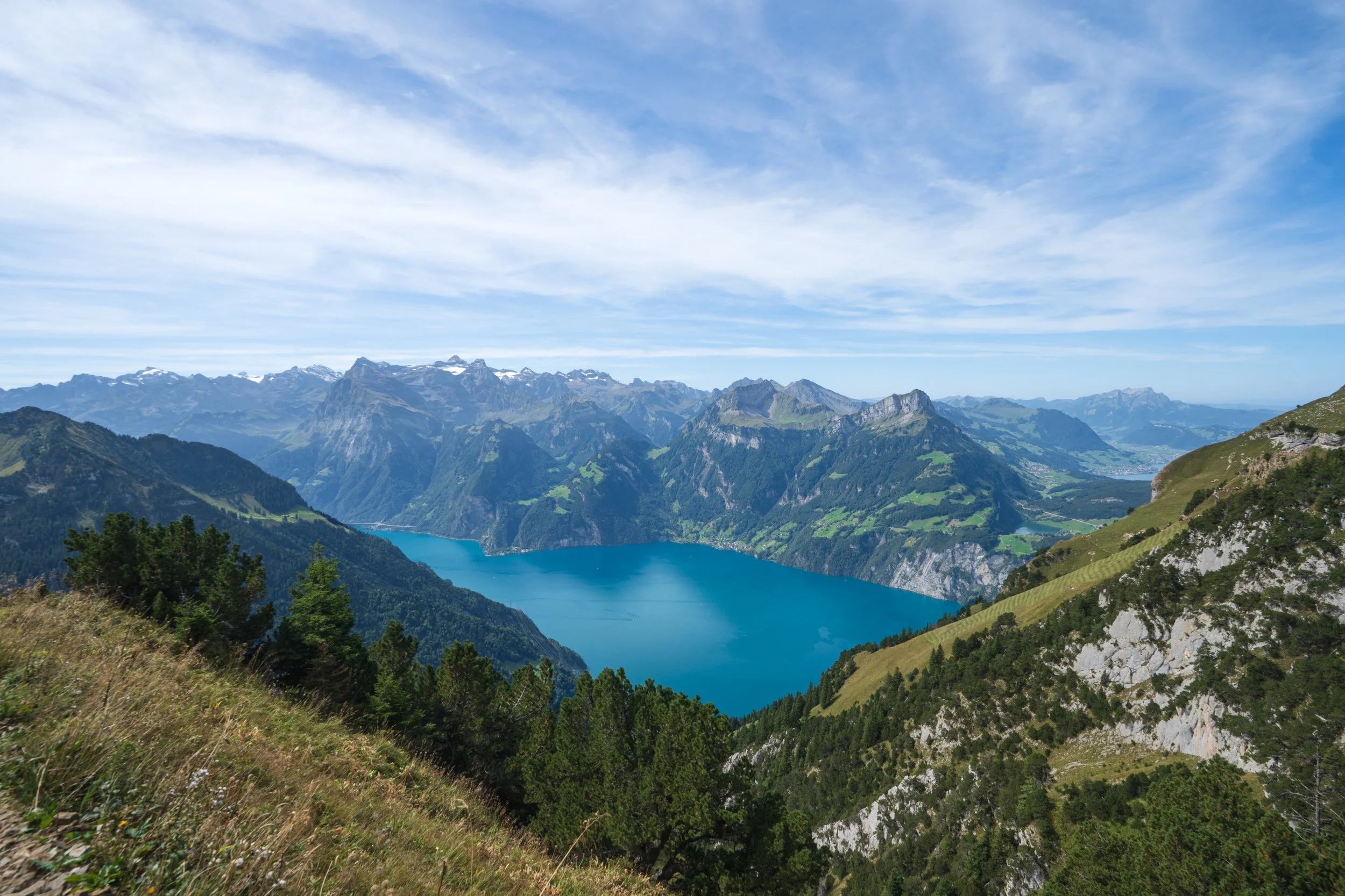 Panoramic landscape featuring snow-capped mountains, rolling green hills, and a blue alpine lake beneath dramatic clouds in the American West.