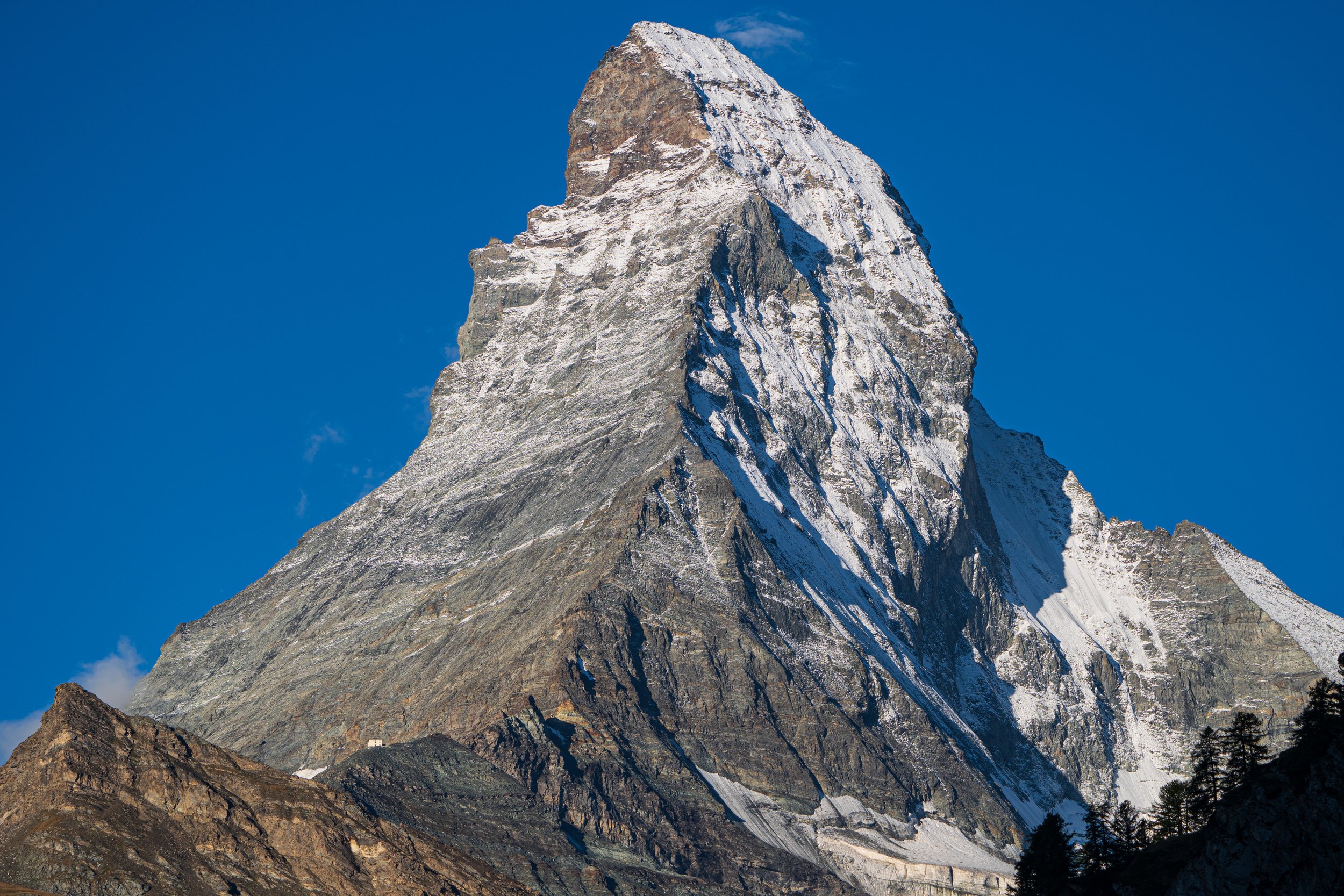 A towering snow-capped mountain peak against a blue sky, with rocky slopes and some trees at the base.