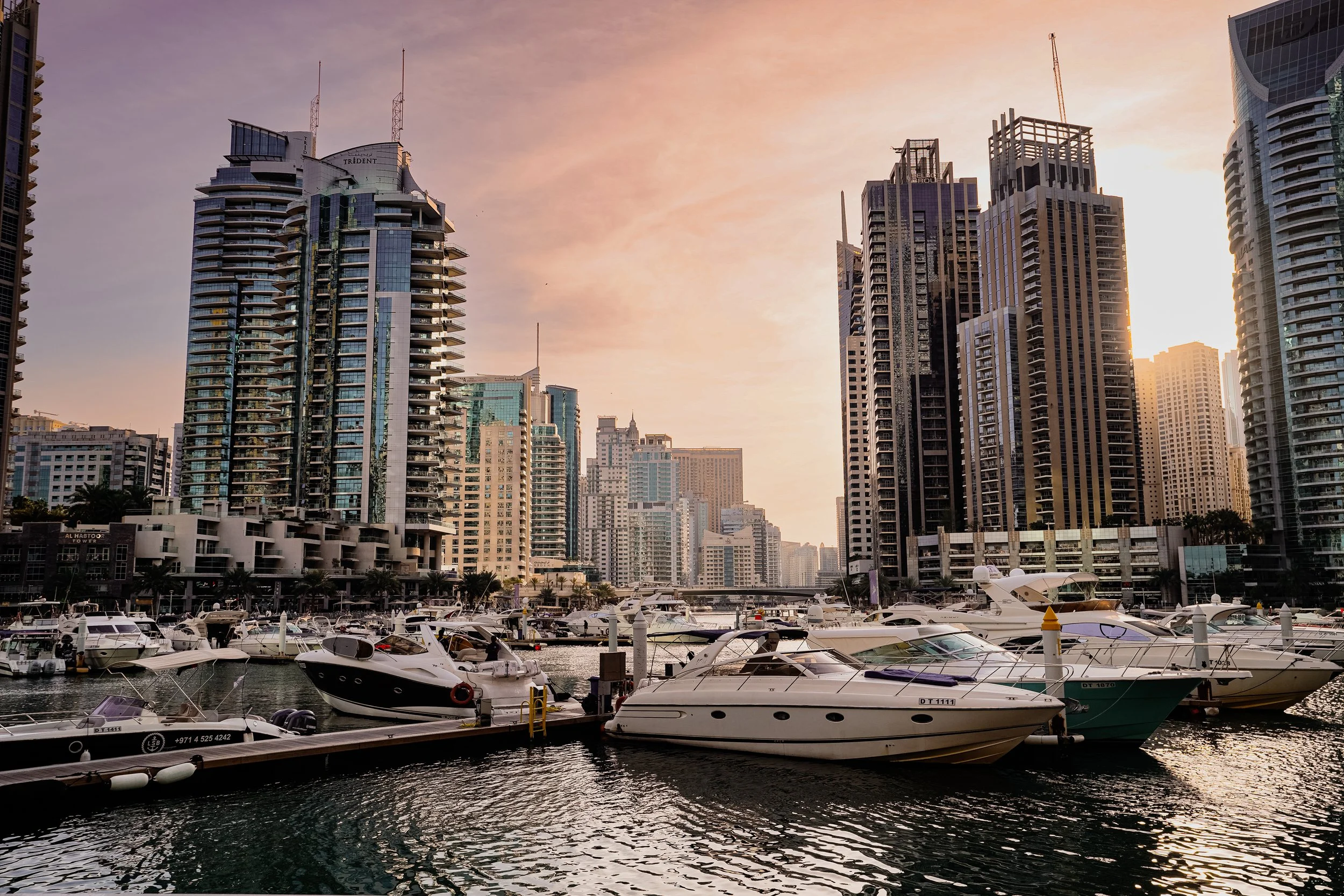 Sunset over a marina with numerous boats docked, surrounded by tall modern skyscrapers in a city skyline.