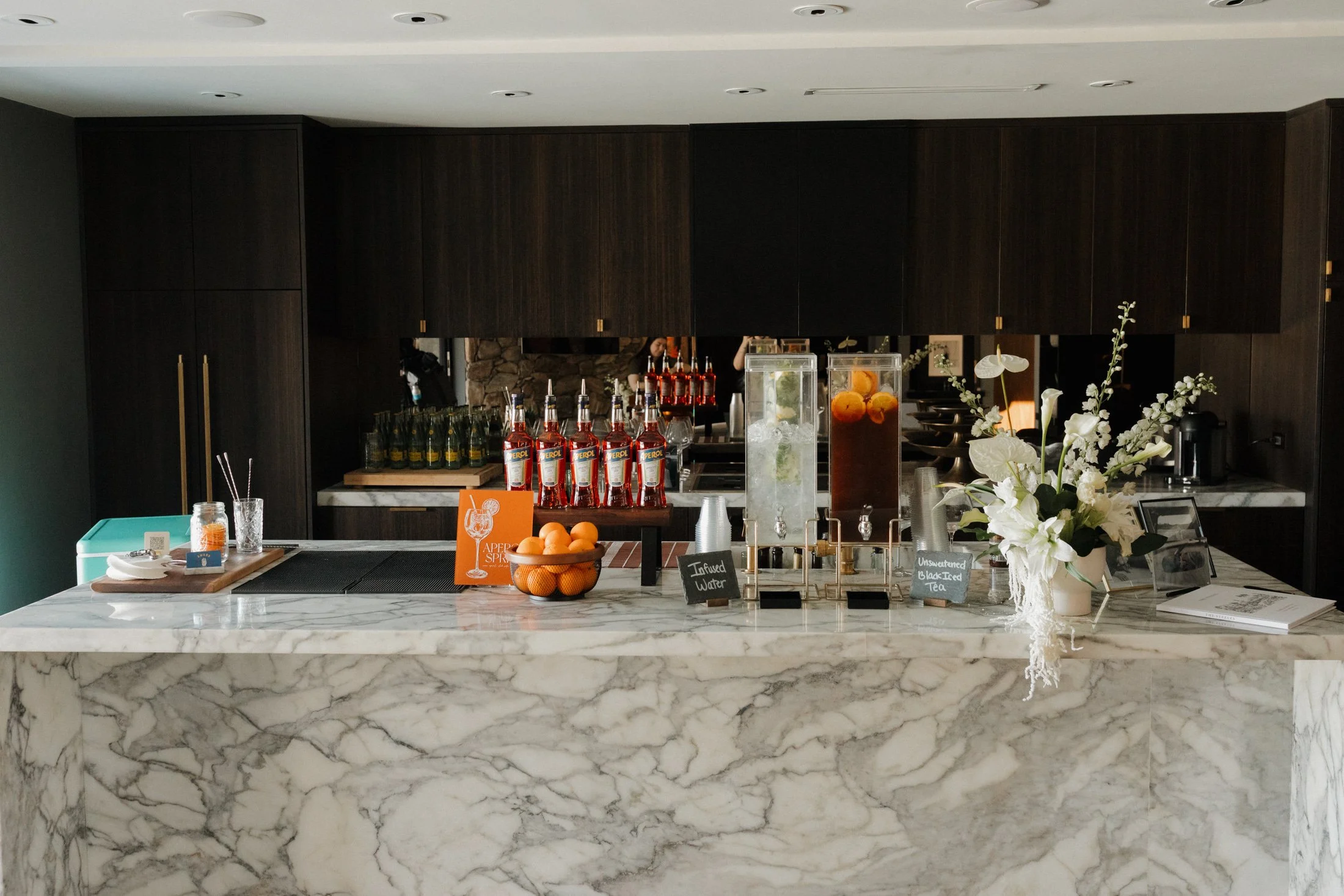 A modern bar area with a marble countertop, dark wood cabinetry, and beverage dispensers including infused water and iced tea, a bowl of oranges, a vase with white flowers, and various bottles and framed pictures.